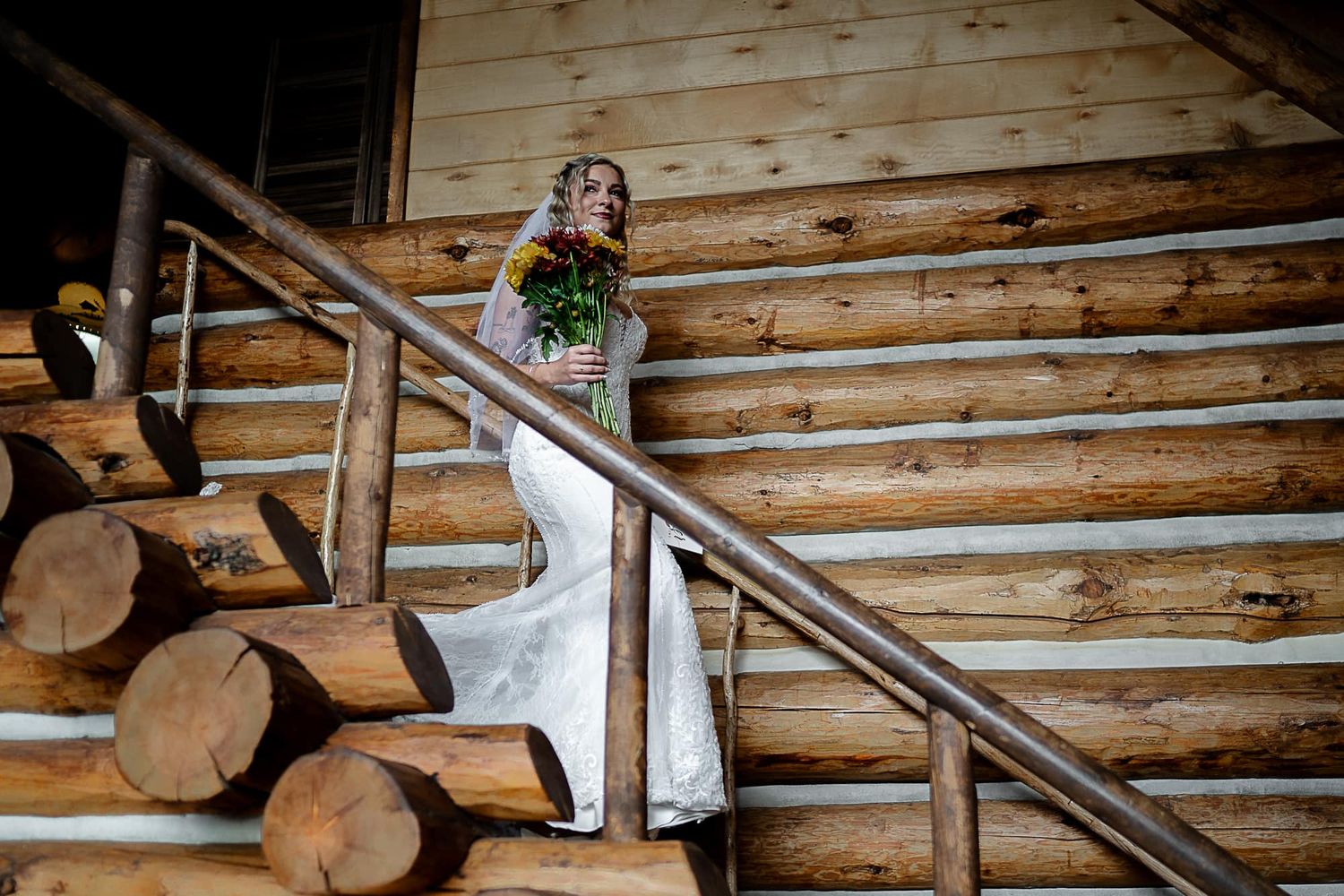 A bride in a white gown stands on rustic wooden stairs with log cabin walls in the background.