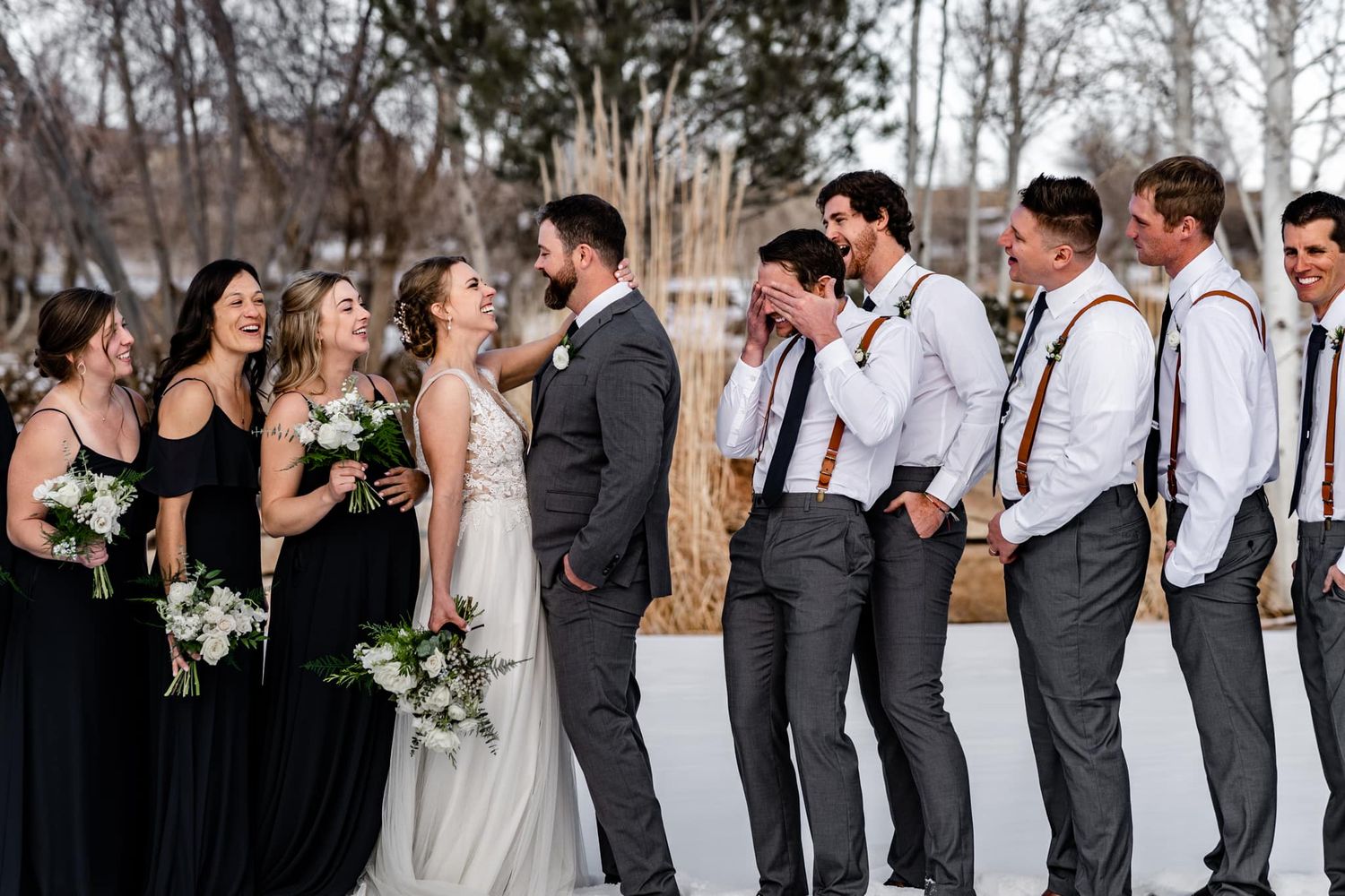 Wedding party in black dresses and suspenders posed in snowy winter setting.