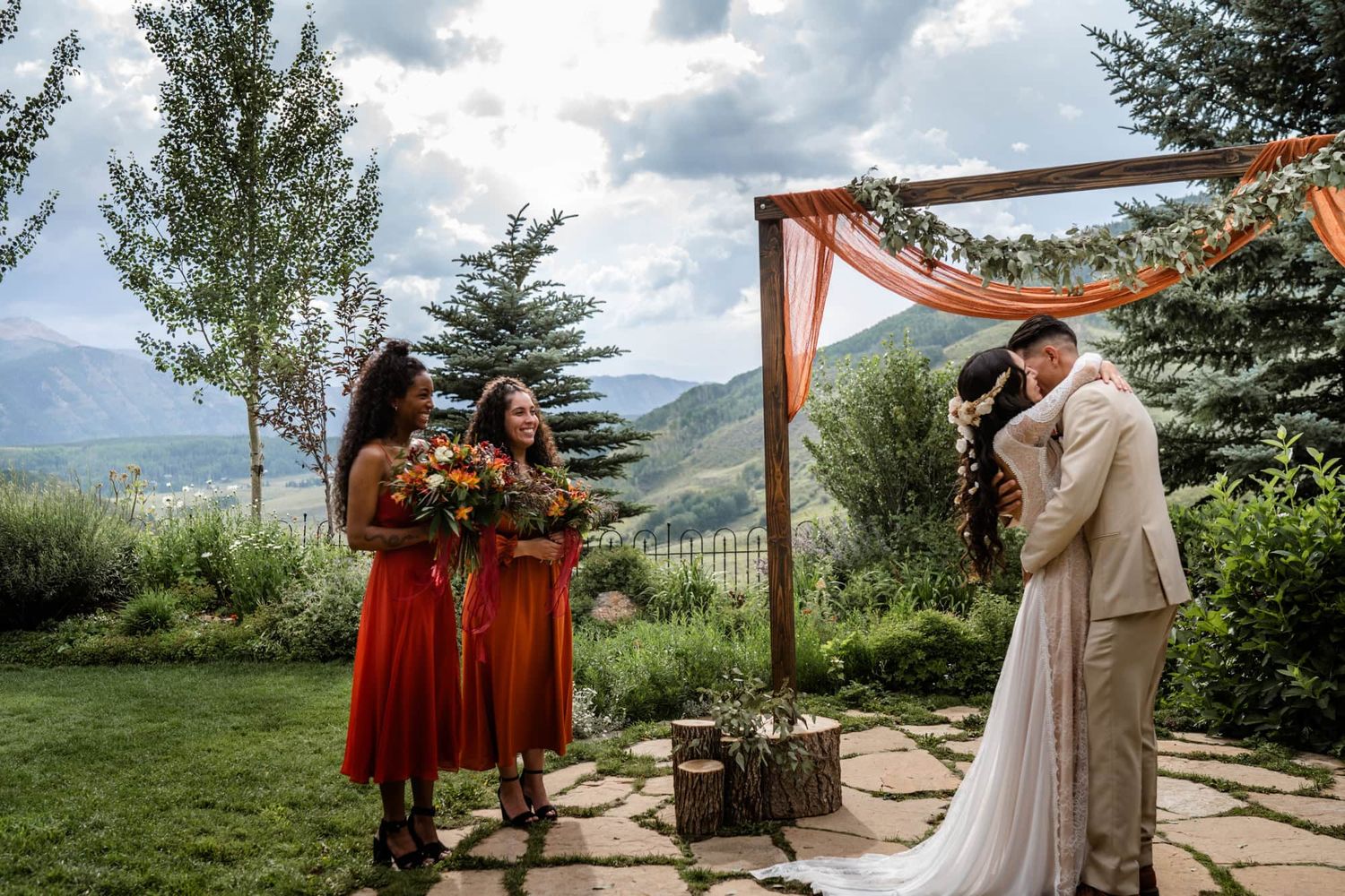 An outdoor wedding ceremony takes place under a wooden arch with mountains and evergreen trees in the background.