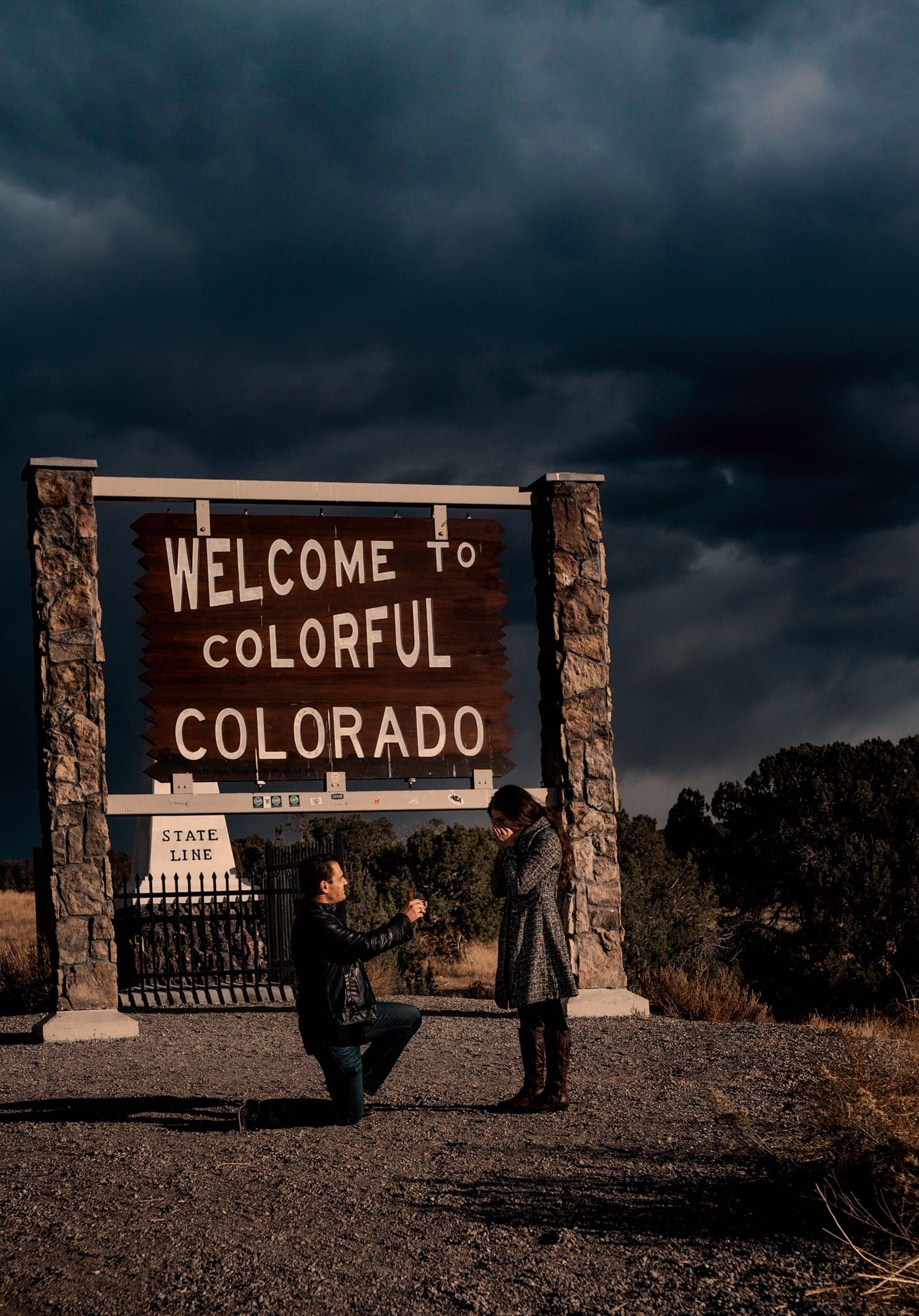 Two people pose at the Welcome to Colorful Colorado sign under stormy skies.