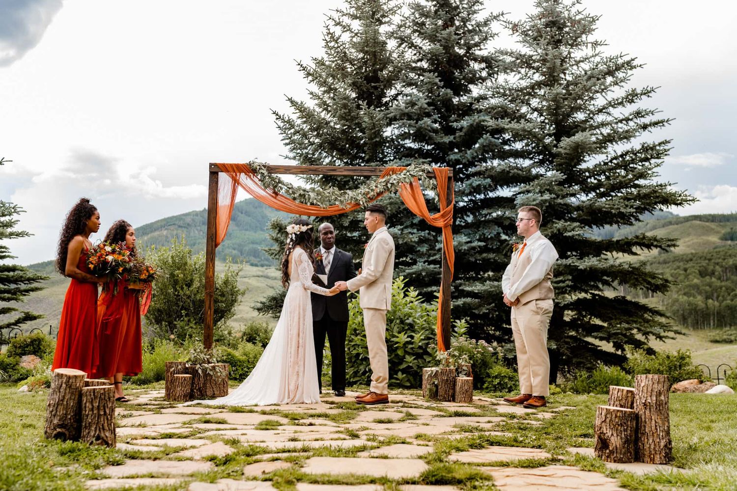 Wedding ceremony with couple at rustic wooden arch surrounded by pine trees and red-dressed bridesmaids.