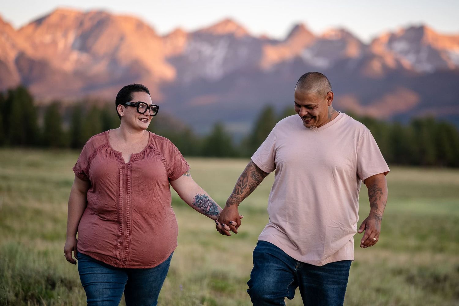 A couple holds hands while walking through a meadow with snow-capped mountains in the background at sunset.