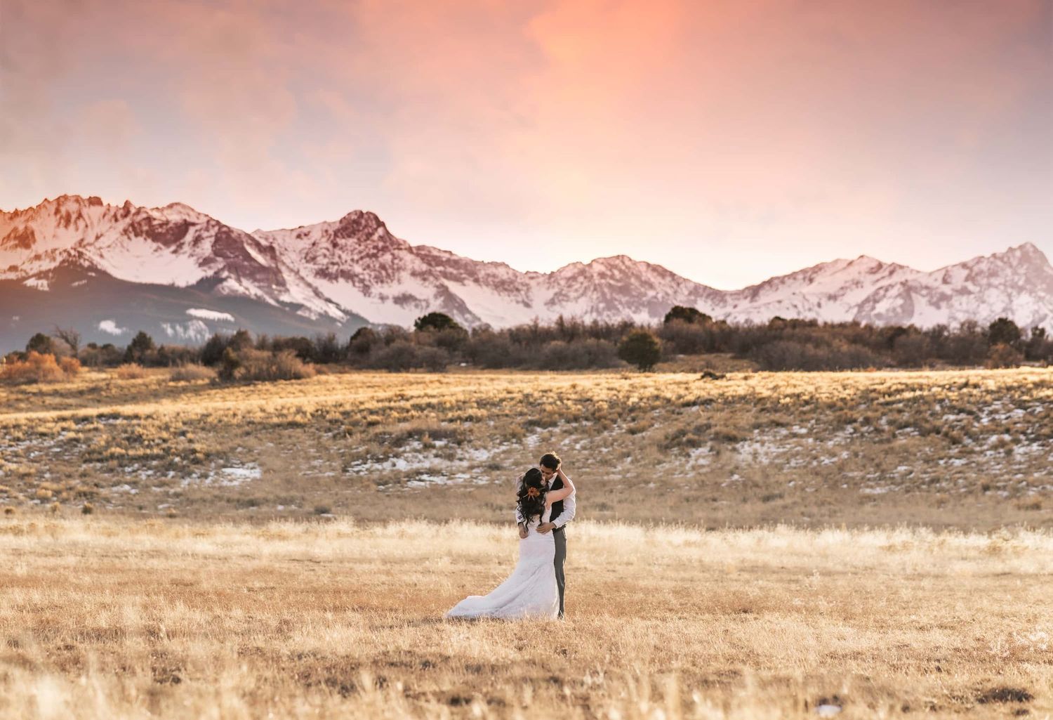 Couple in wedding attire standing in a golden field with snow-capped mountains at sunset.