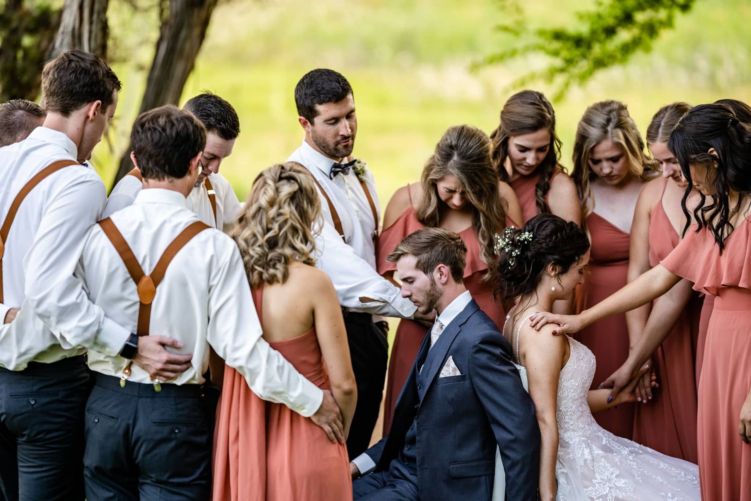 Wedding party with bridesmaids in pink dresses and groomsmen in suspenders gathered in a circle outdoors.