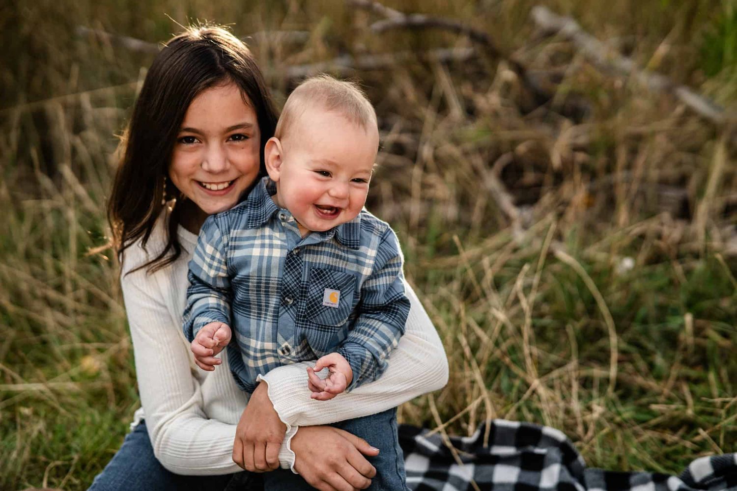 Happy toddler in plaid jacket laughing while being held outdoors in tall grass.