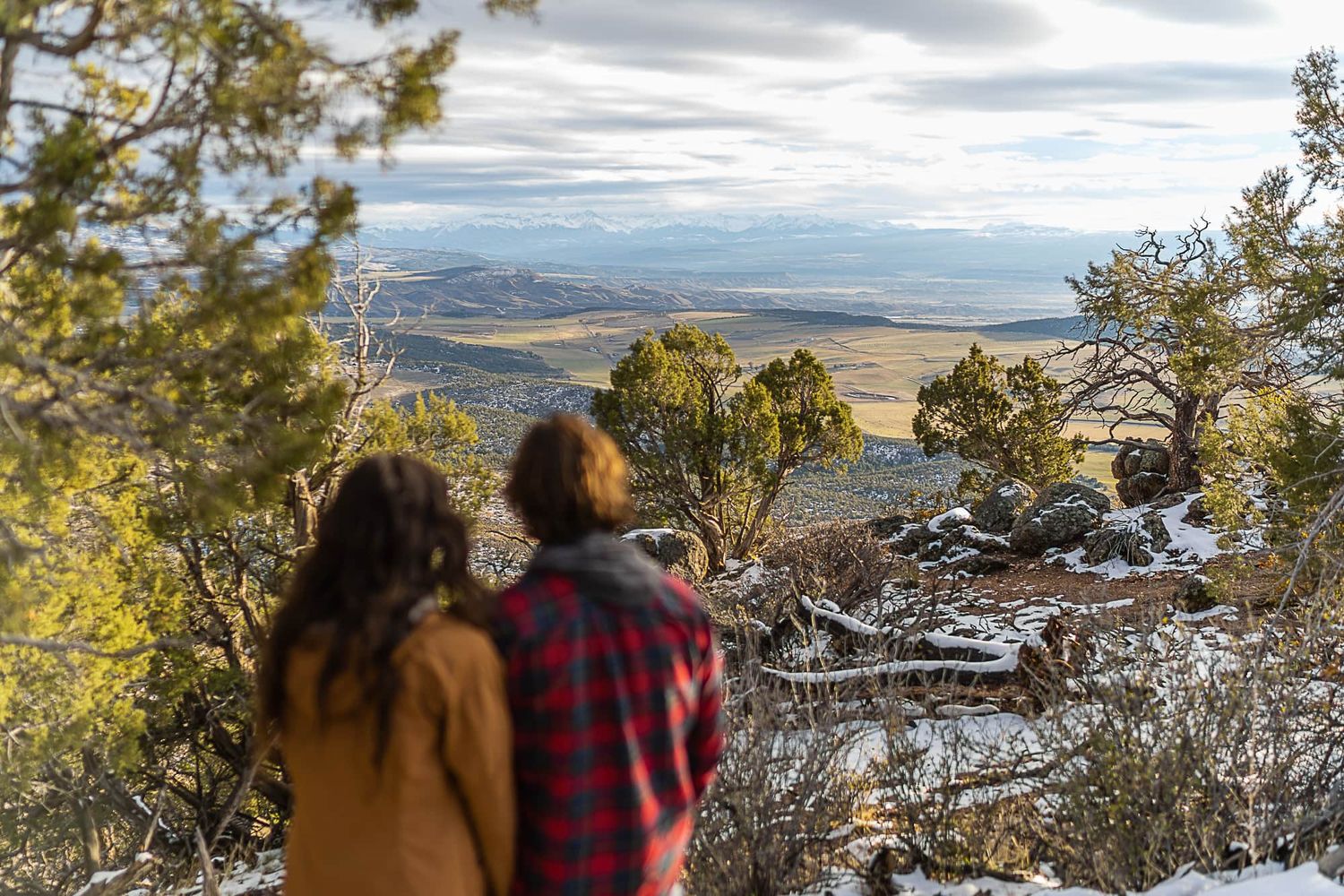 A couple admires the scenic mountain valley view from a snow-covered overlook.