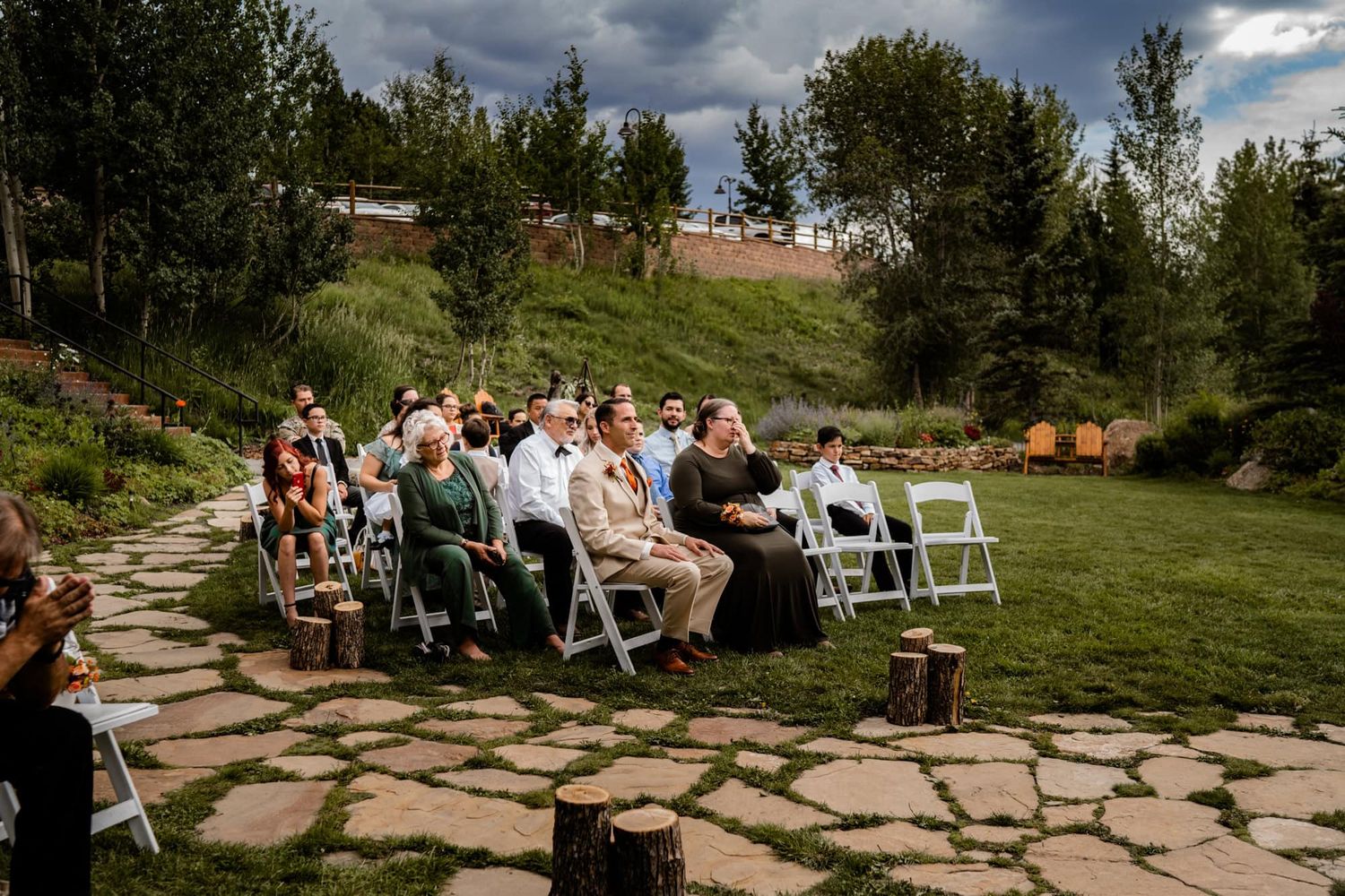 Outdoor wedding ceremony guests seated on white chairs on a stone patio surrounded by green landscaping under cloudy skies.