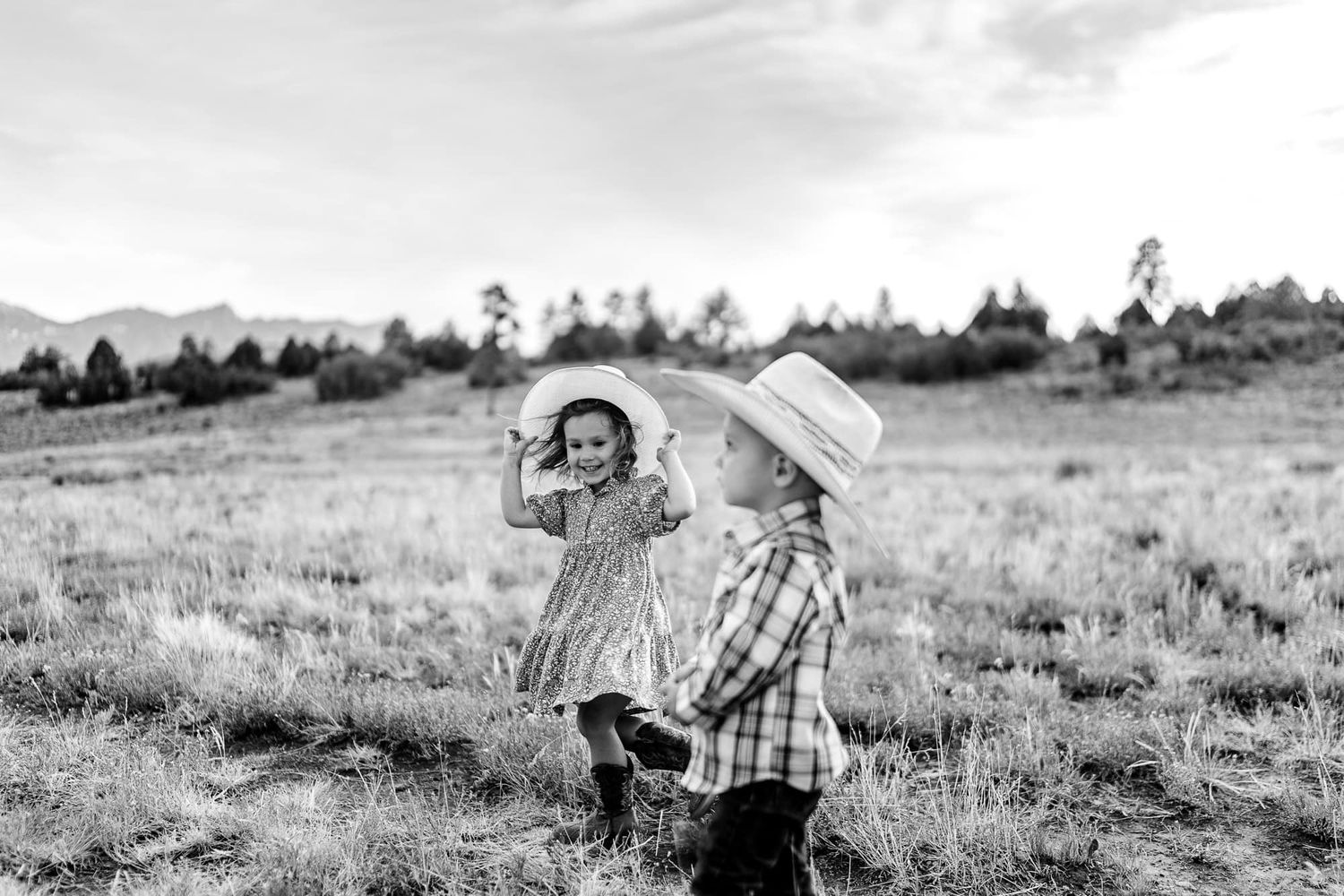Two children in western attire play together in a field with mountains in the background.