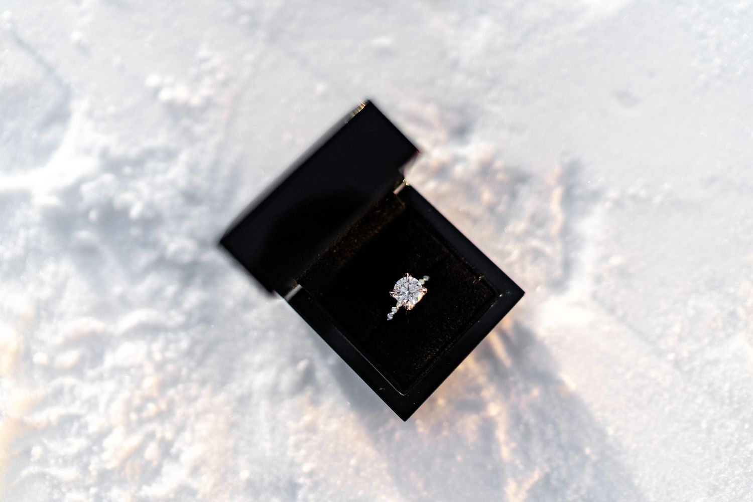 Diamond engagement ring in black box photographed on snowy surface.