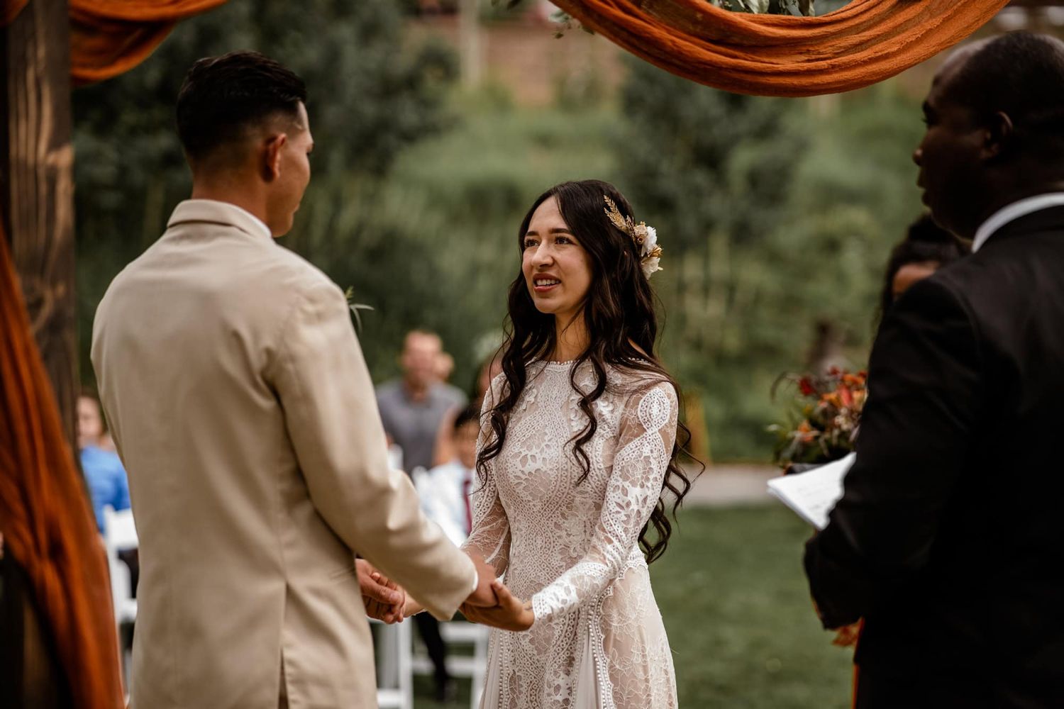 Couple stands at altar under draped orange fabric during outdoor forest wedding ceremony with lace dress and tan suit.