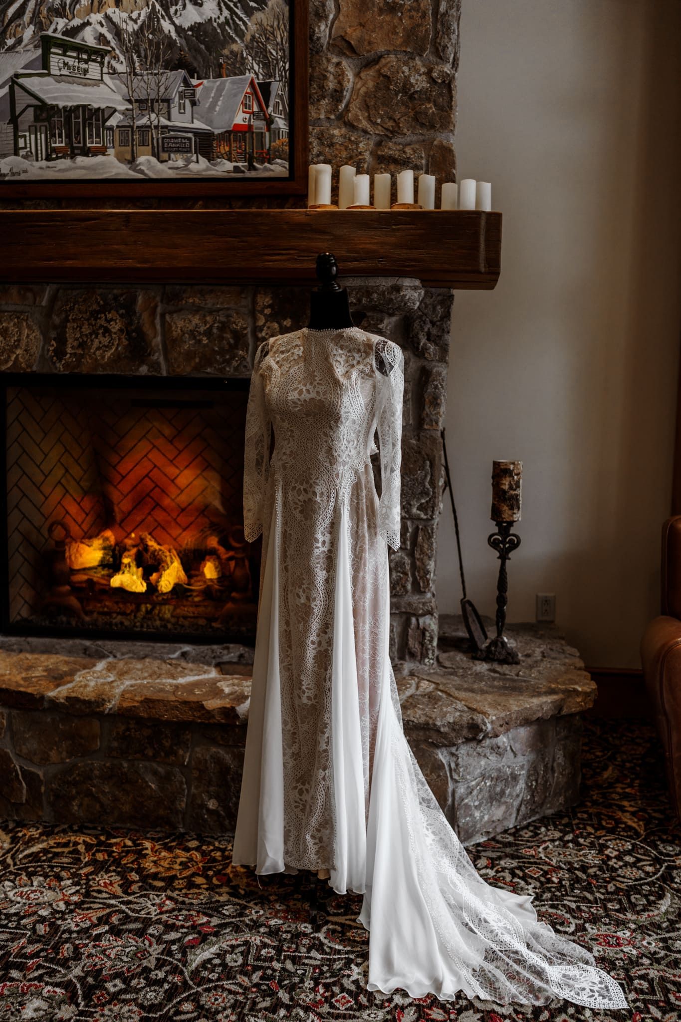 Elegant white lace wedding dress displayed in front of cozy stone fireplace with glowing flames and rustic mantel decor.