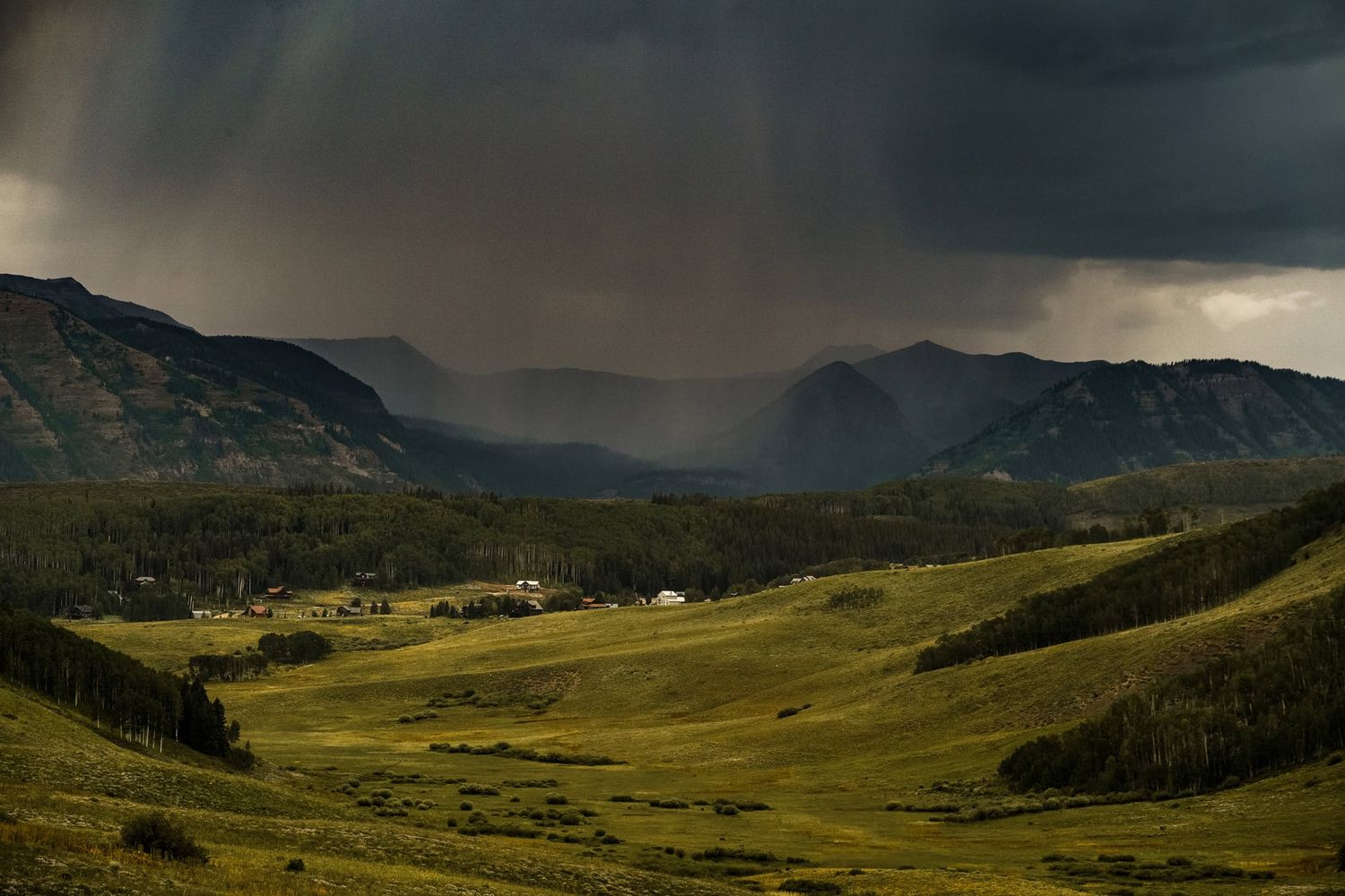 Storm clouds gather over green mountain meadows in a dramatic landscape with rays of sunlight breaking through.