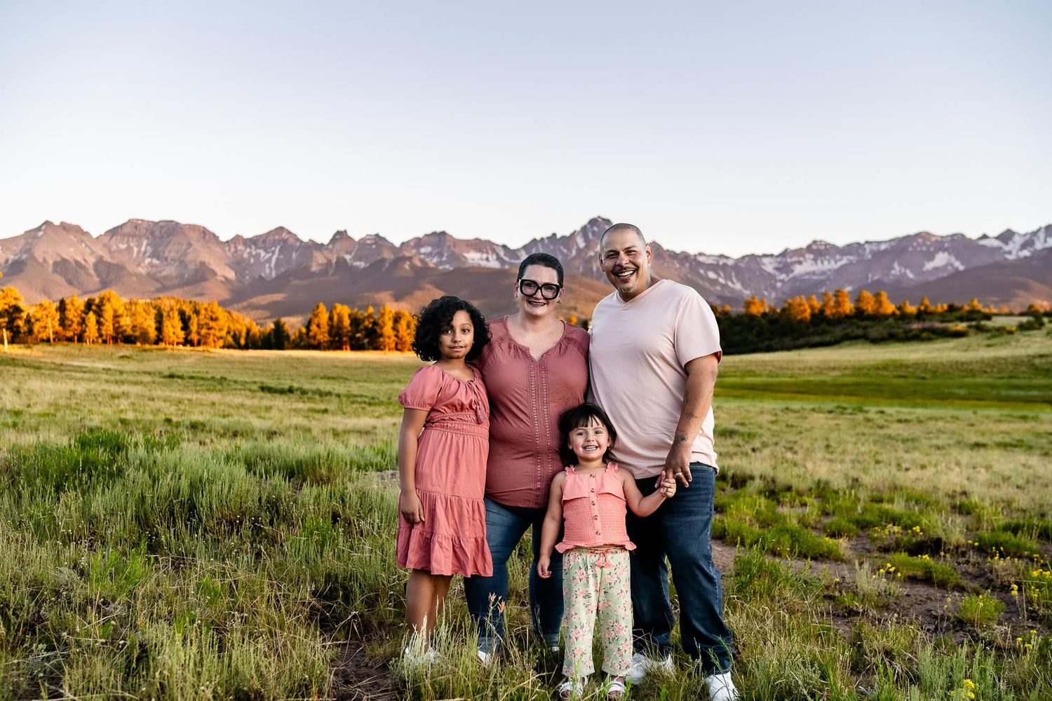 A family poses together in a meadow with snow-capped mountains in the background. Captured by Ridgway Colorado Family Photographer.