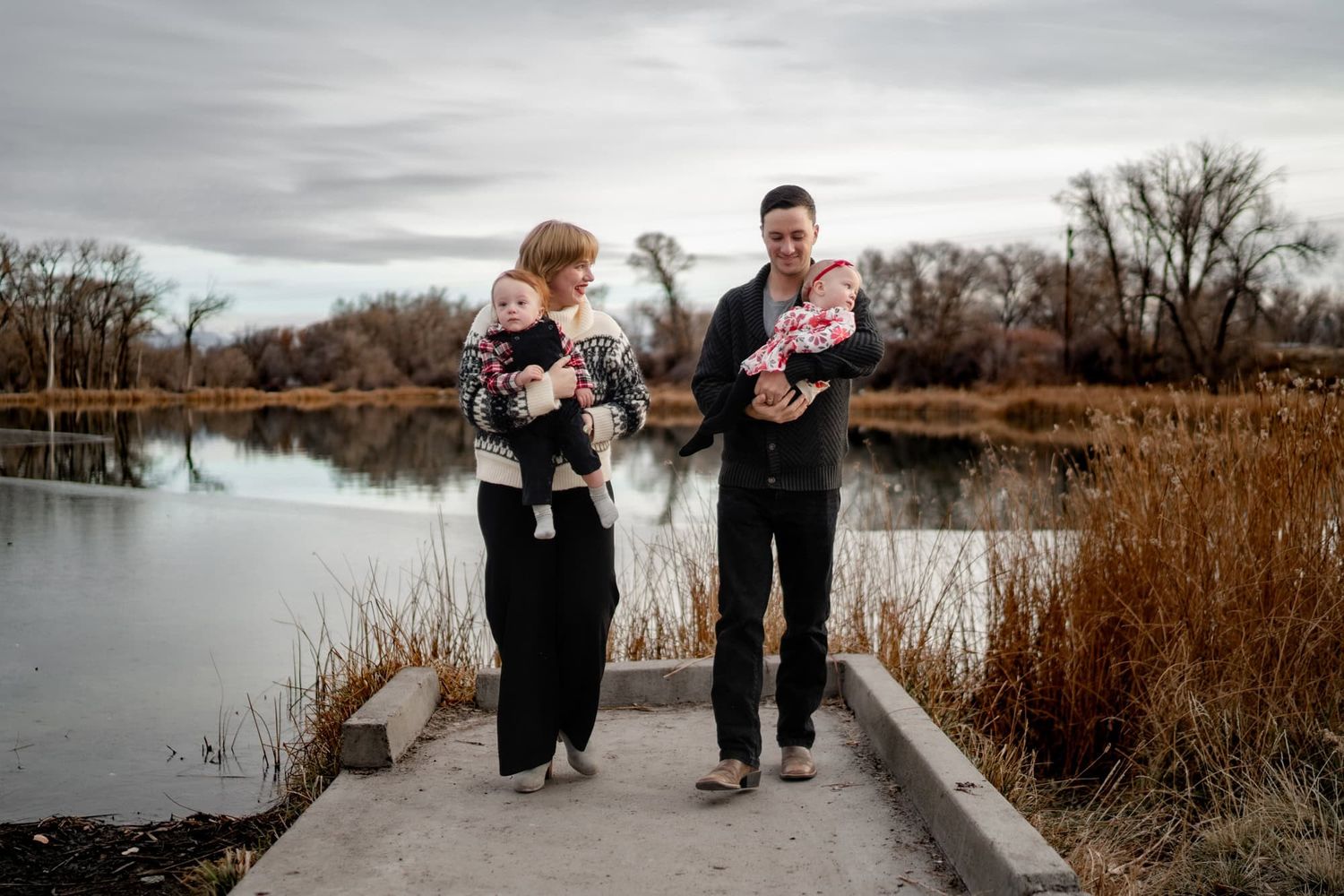 A couple stands on a path near a lake while holding their twin babies.