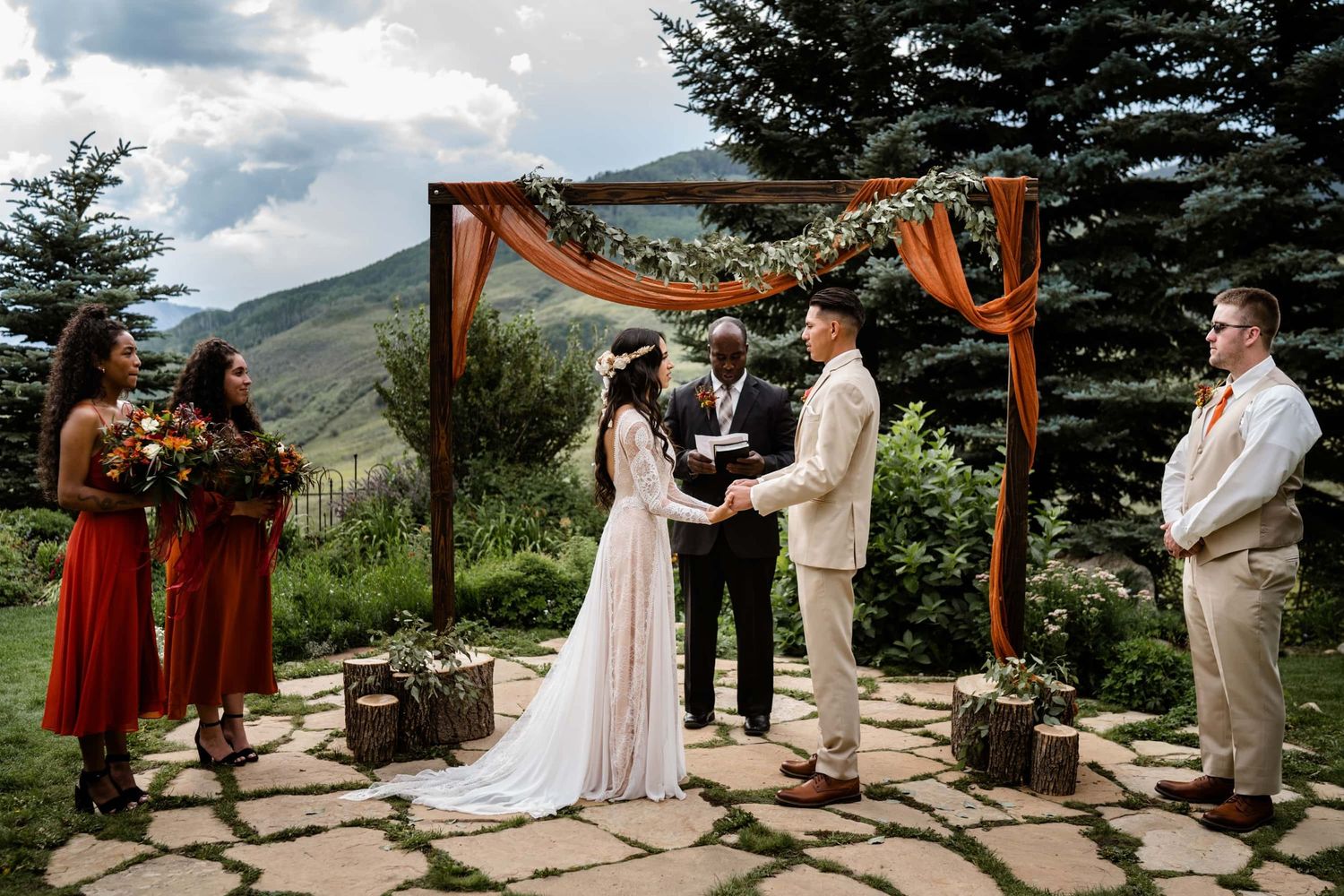 A rustic mountain wedding ceremony takes place under a wooden arch decorated with fabric on a stone patio surrounded by evergreens.
