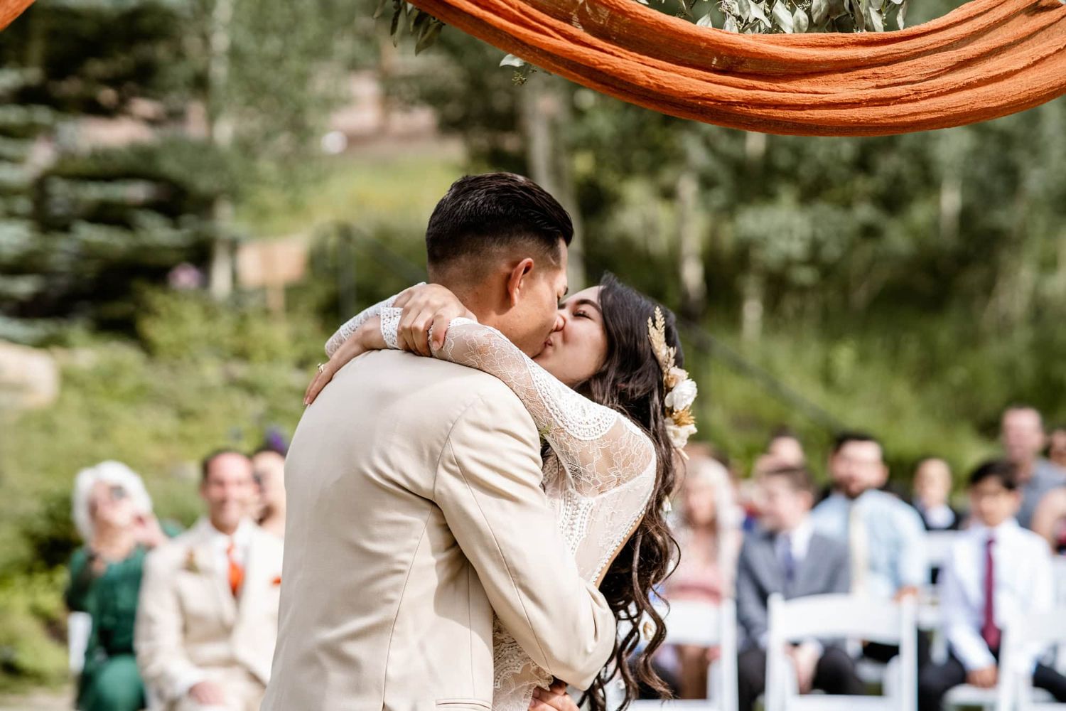 A couple shares a passionate kiss outdoors during their wedding ceremony with guests looking on in the background.