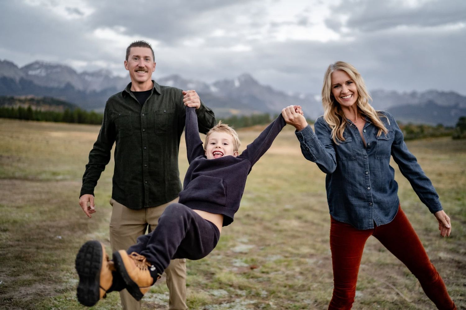 A family plays together outdoors, swinging a child between them with mountains in the background.