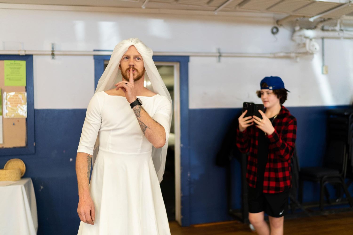 Playful moment in a gymnasium with man in wedding dress making gestures to keep the secret that it's no the bride surprising the groom.