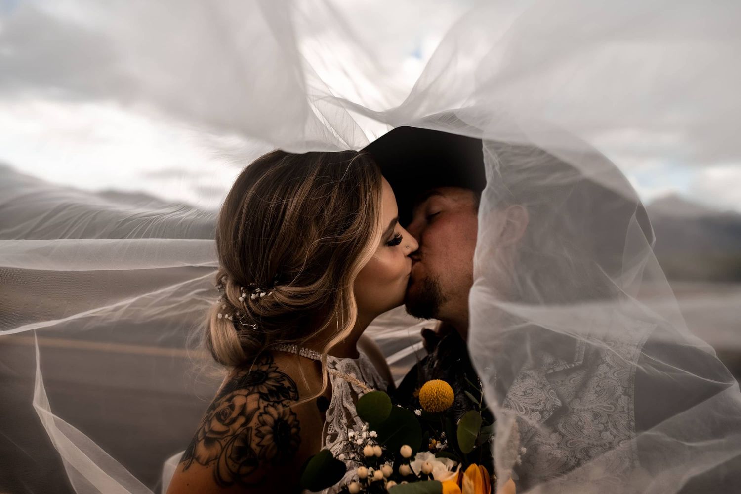 Intimate wedding kiss captured under flowing white veil with dramatic sky backdrop.