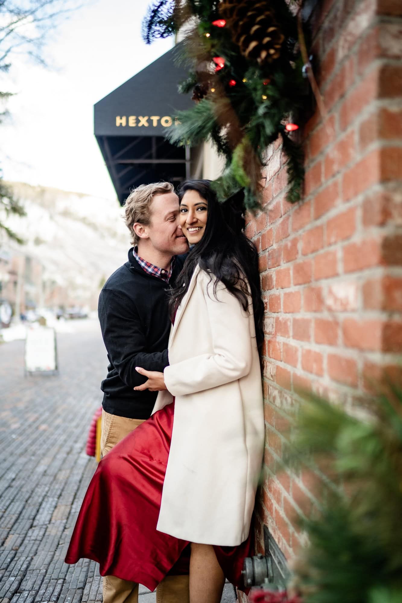 A romantic winter moment with a couple embracing by a brick wall decorated with holiday greenery.