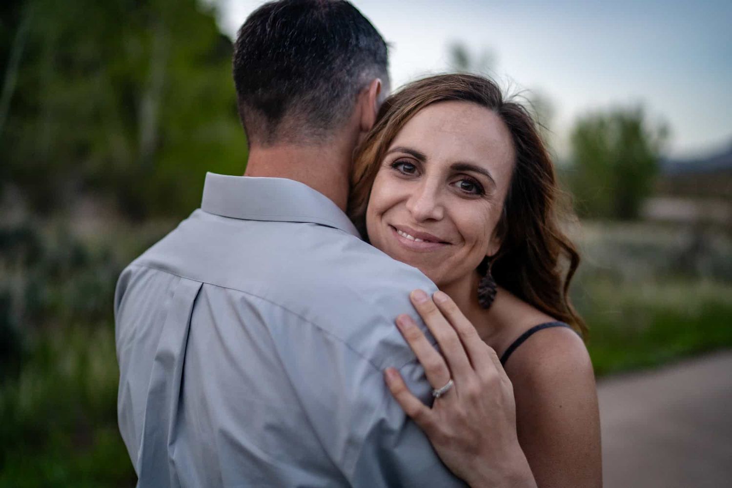 Couple sharing an intimate moment outdoors as the woman embraces her partner while showing off an engagement ring.