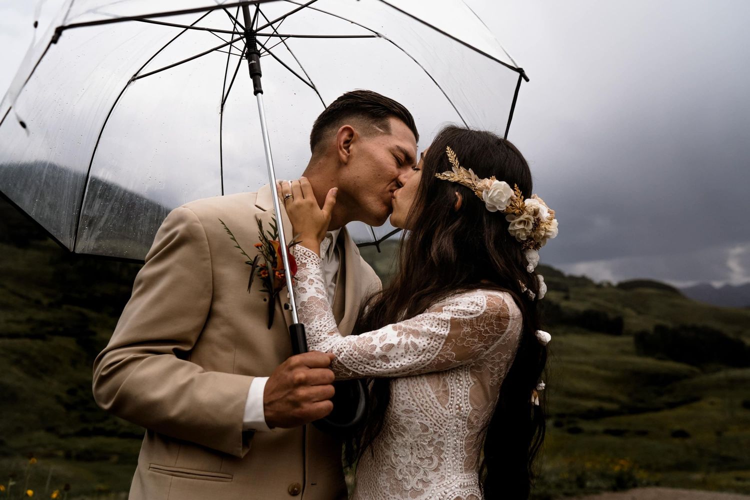 A couple shares an intimate moment under a clear umbrella against a backdrop of moody mountains and stormy skies.