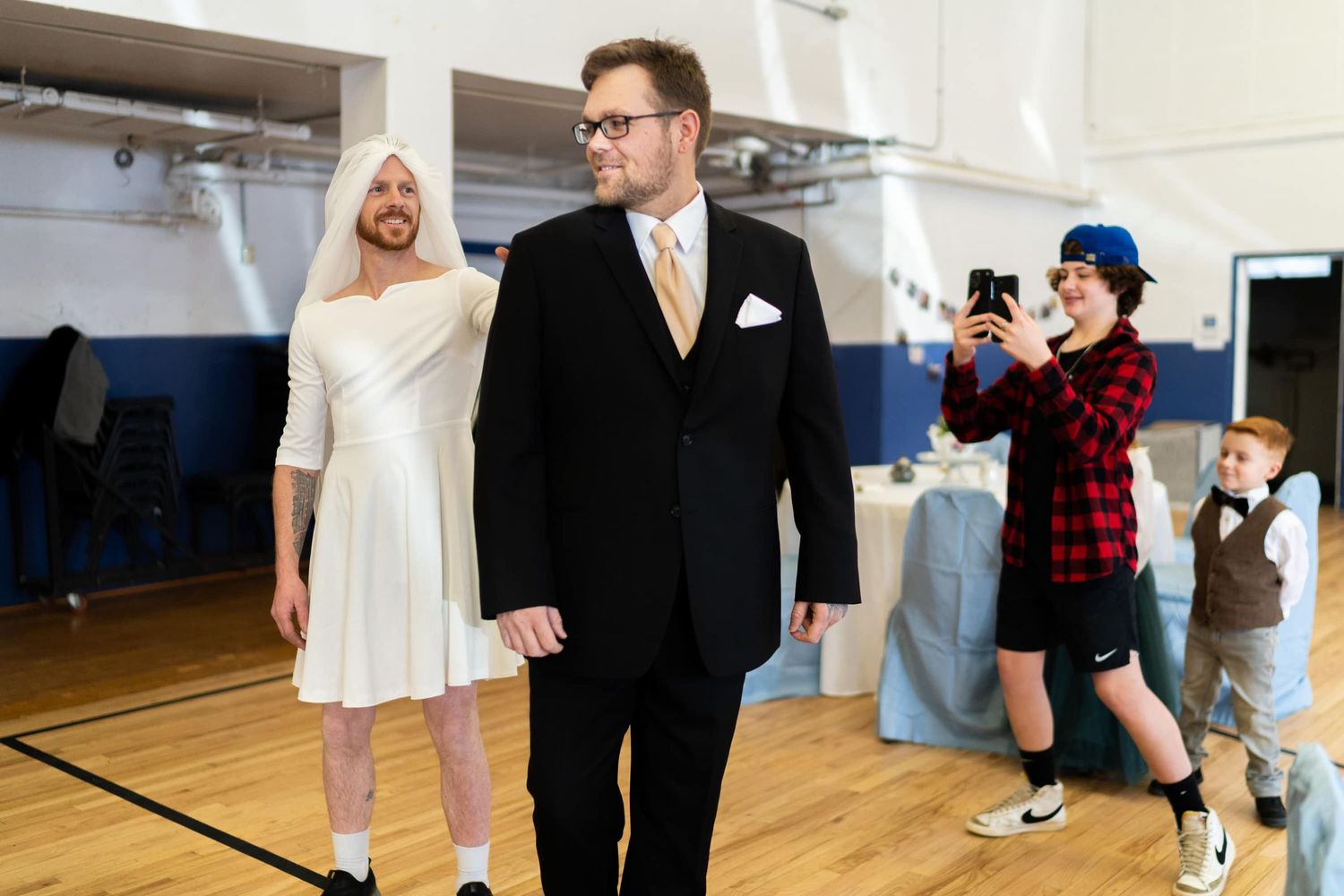 Groom waits for bride in gymnasium while photographer captures the moment. Turns out it's his brother in a dress.