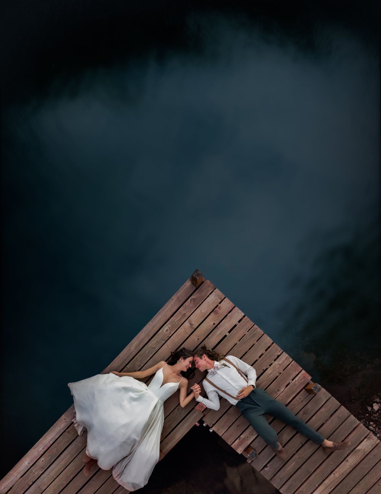 Aerial view of couple lying on wooden dock over dark water at dusk.