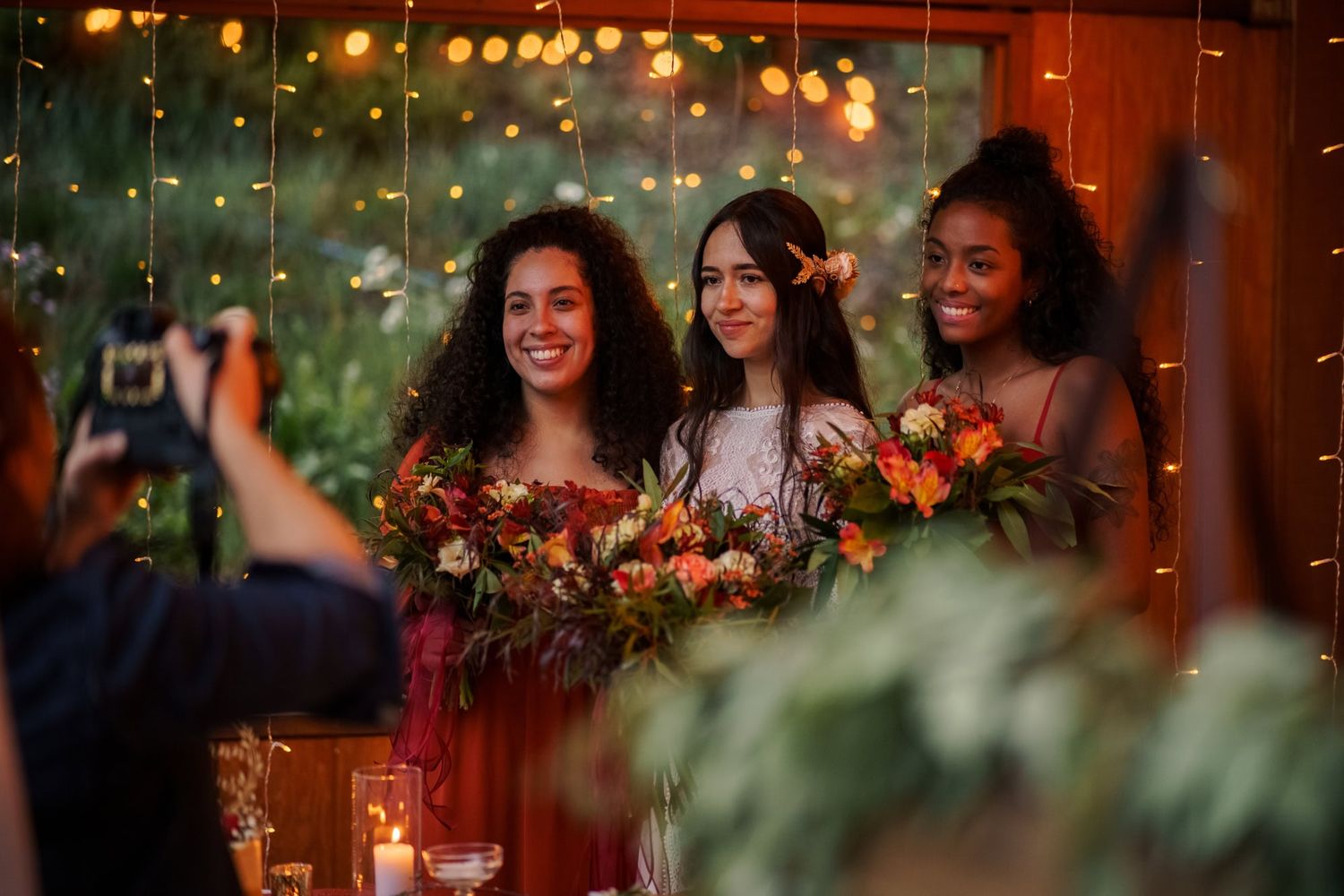 Three bridesmaids in elegant dresses hold bouquets while smiling during an evening wedding reception with twinkle lights.