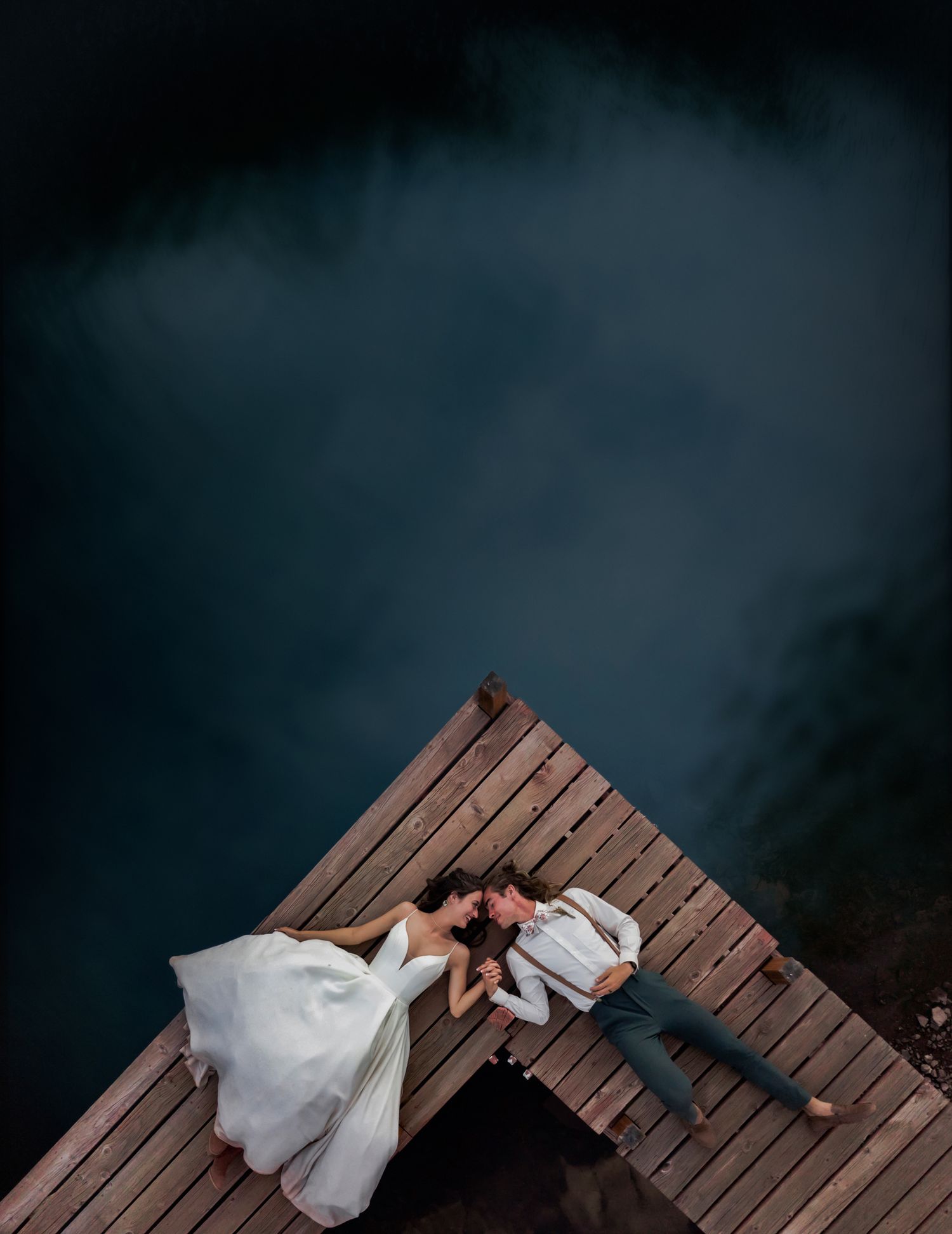 Aerial view of couple lying on wooden dock over dark water in romantic wedding photo.