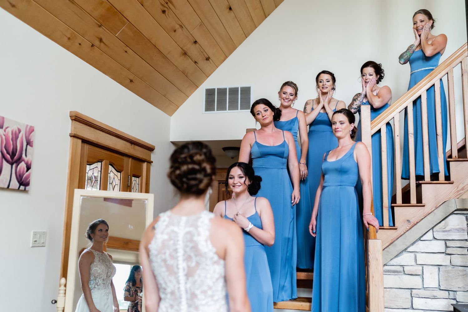 Bridesmaids in matching blue dresses react with joy on a staircase to seeing the bride.