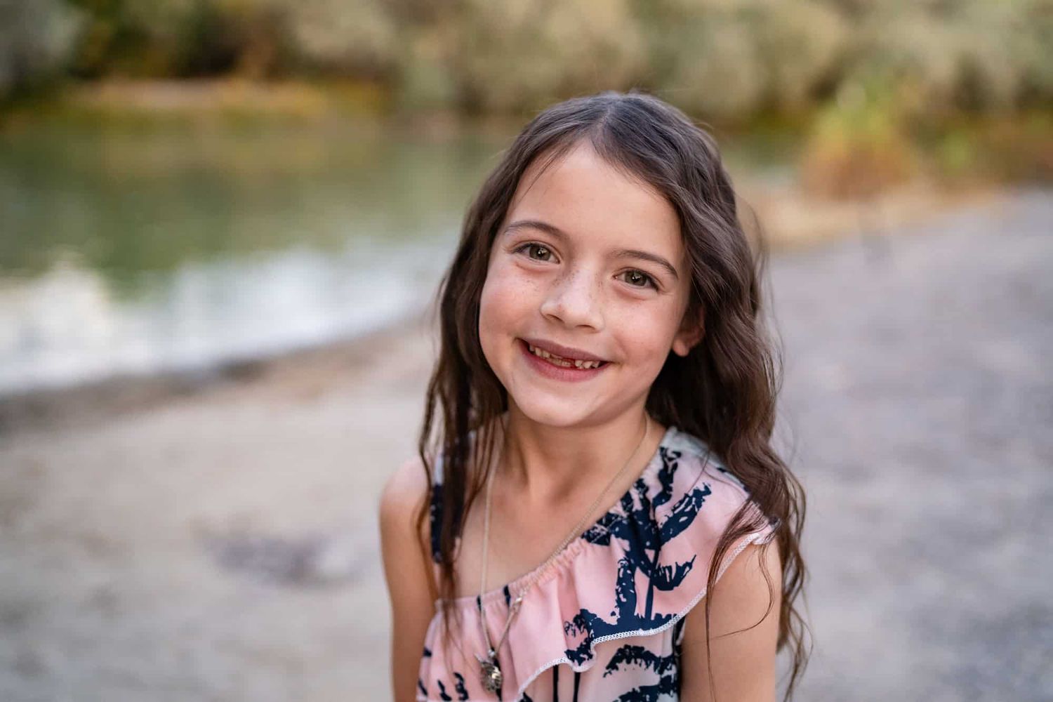 Young child smiling brightly outdoors near a body of water.