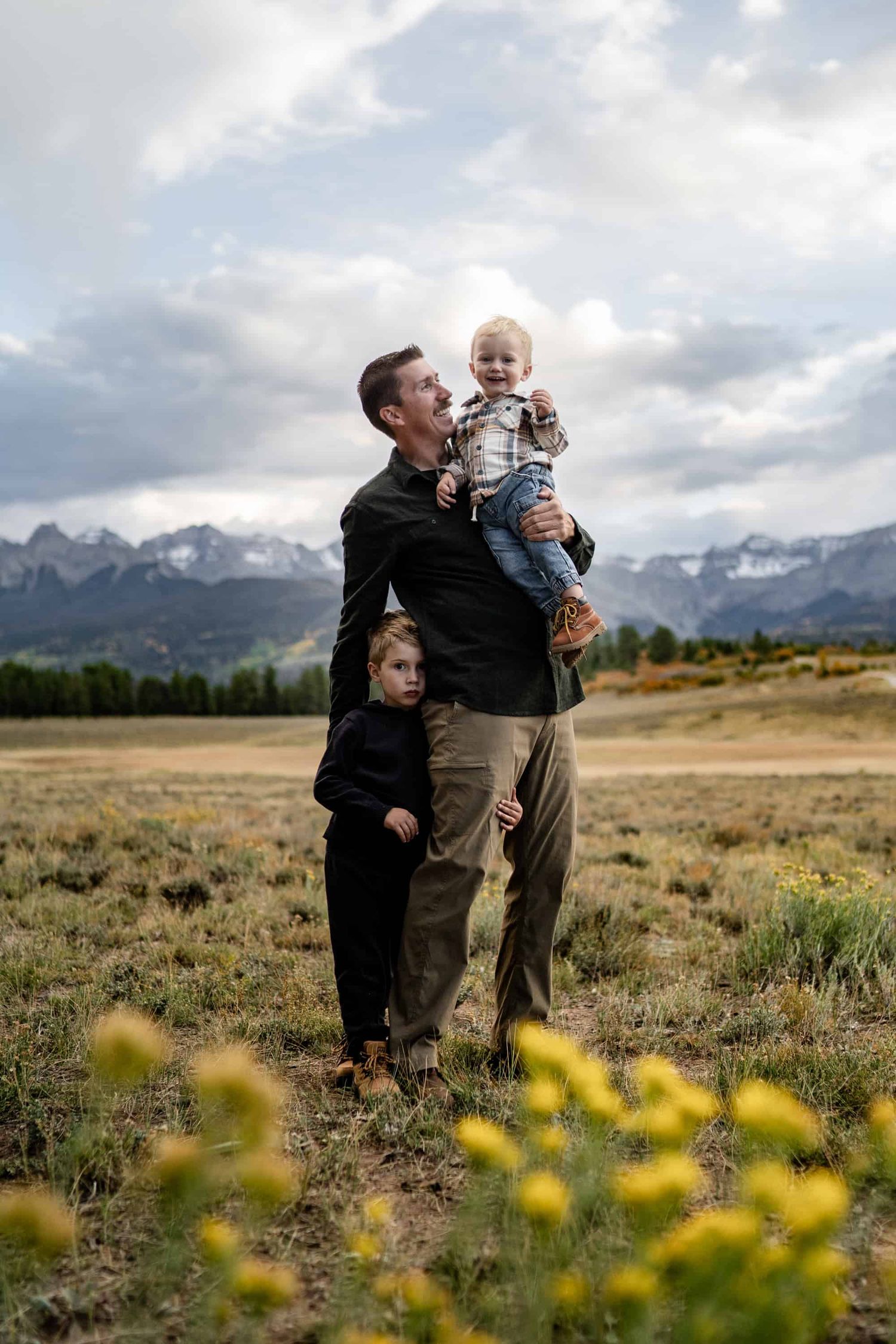 Parent and children sharing playful moment in mountain meadow with yellow wildflowers. Best place to take family portraits in Colorado.