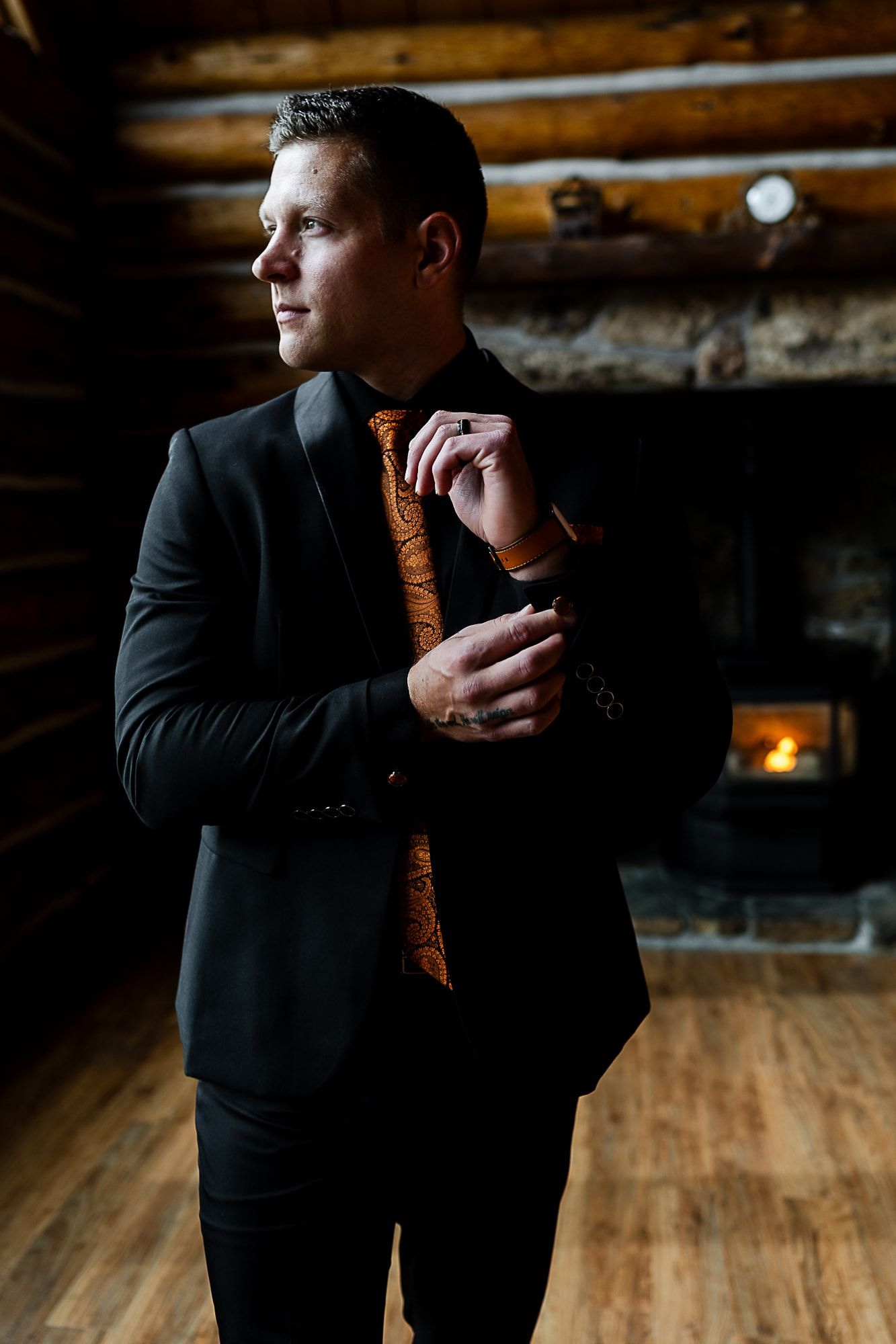 Groom in black suit with orange tie adjusts cufflinks against wooden cabin interior.