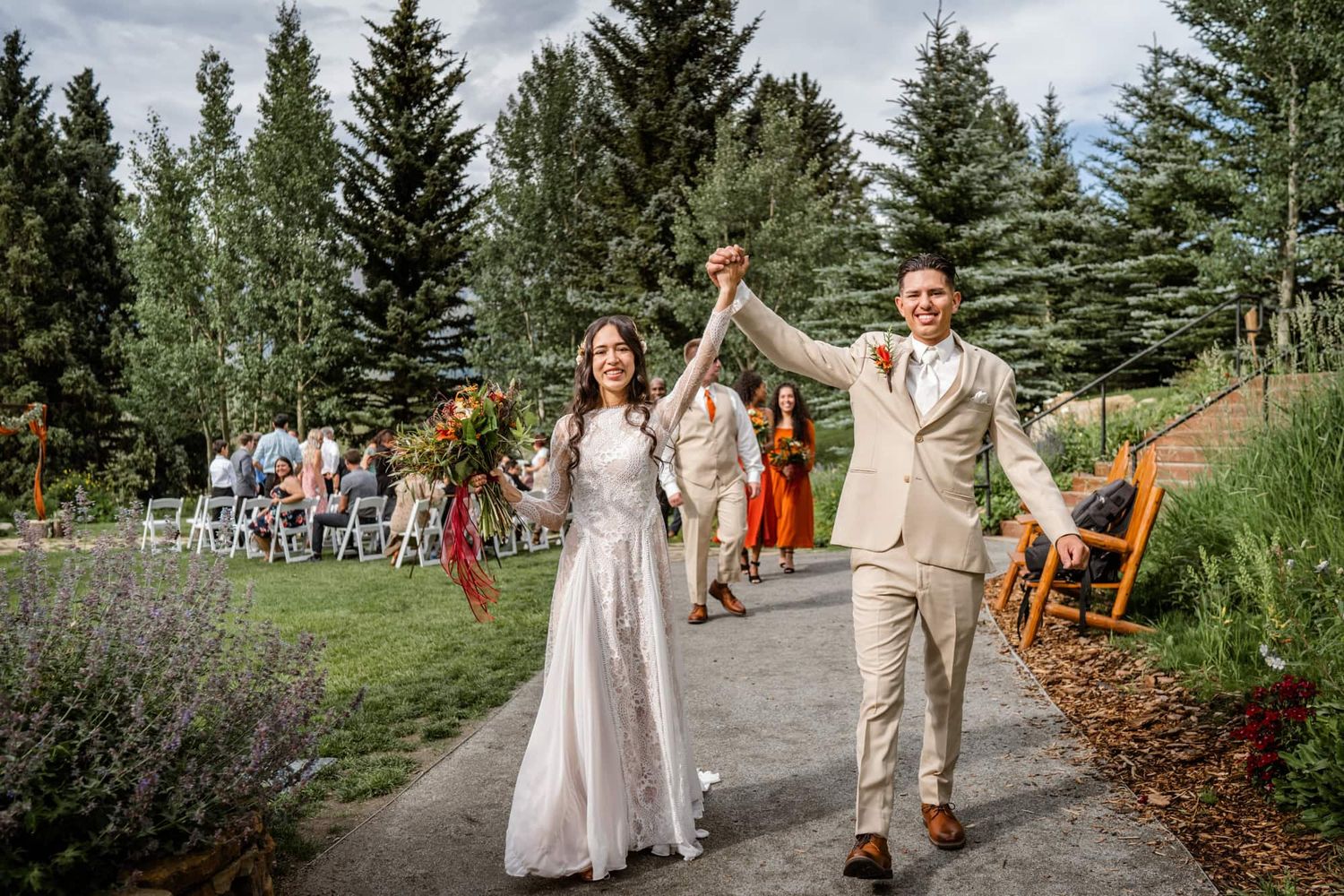 A couple celebrating after their wedding ceremony on a path surrounded by evergreen trees and lavender flowers.