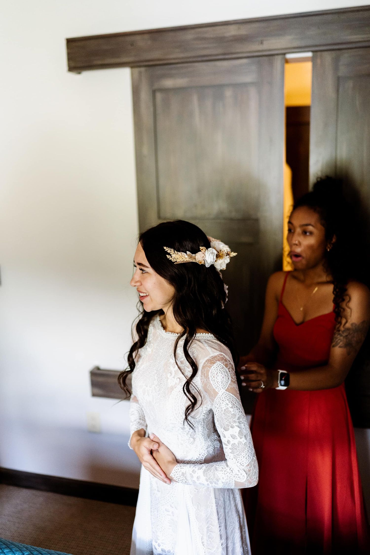 Two people in a room, one wearing a delicate white lace wedding dress with long sleeves and another in a red dress.
