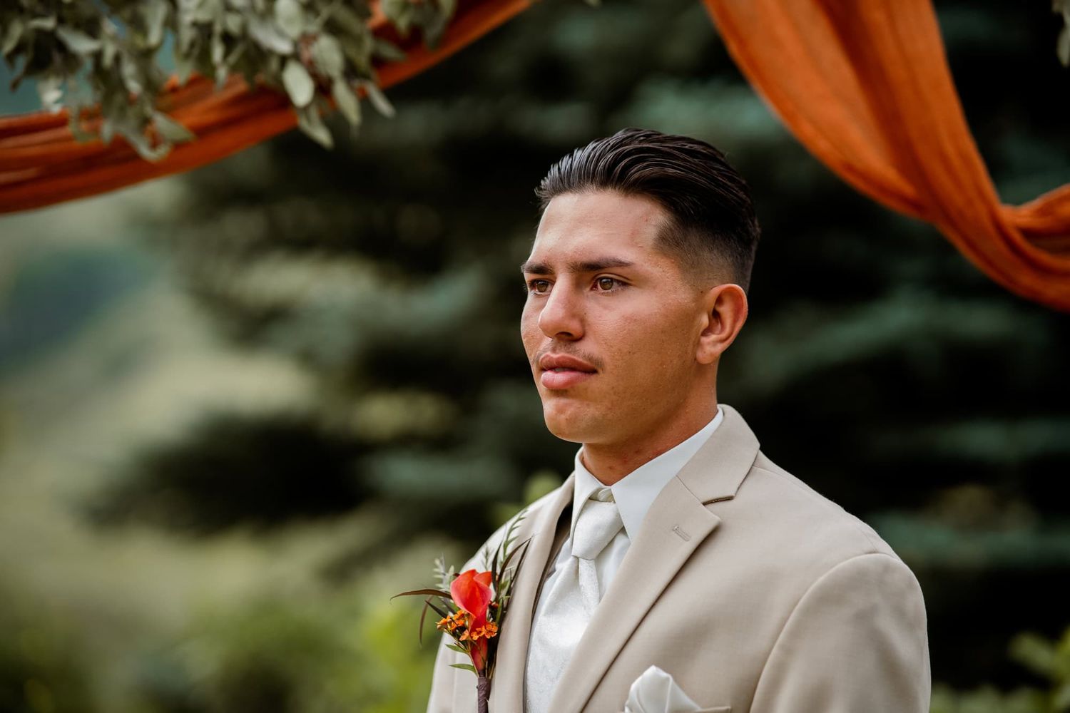 A groom in a light beige suit and white tie stands under orange and green outdoor wedding decor.