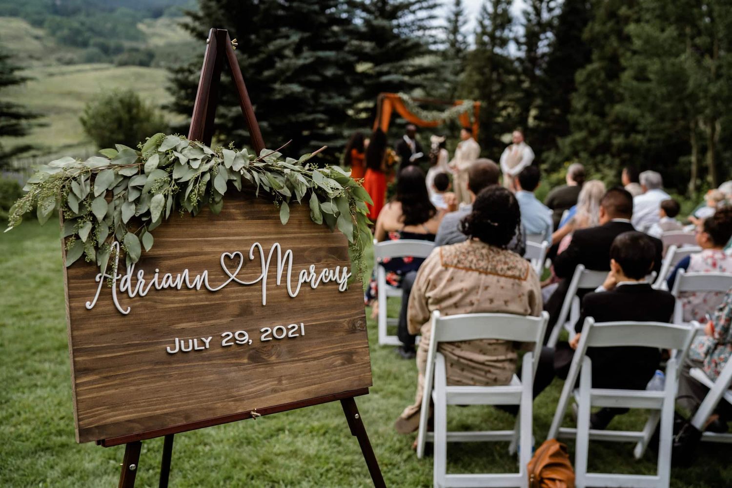 Wooden wedding welcome sign with greenery and guest seating at mountain outdoor ceremony venue.