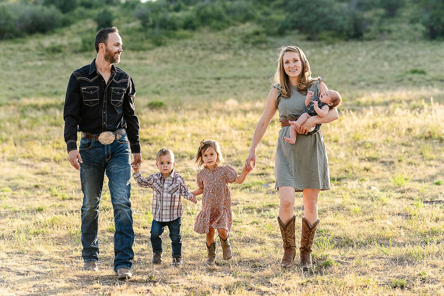 Family of four walking together through a golden meadow with mountains in background.