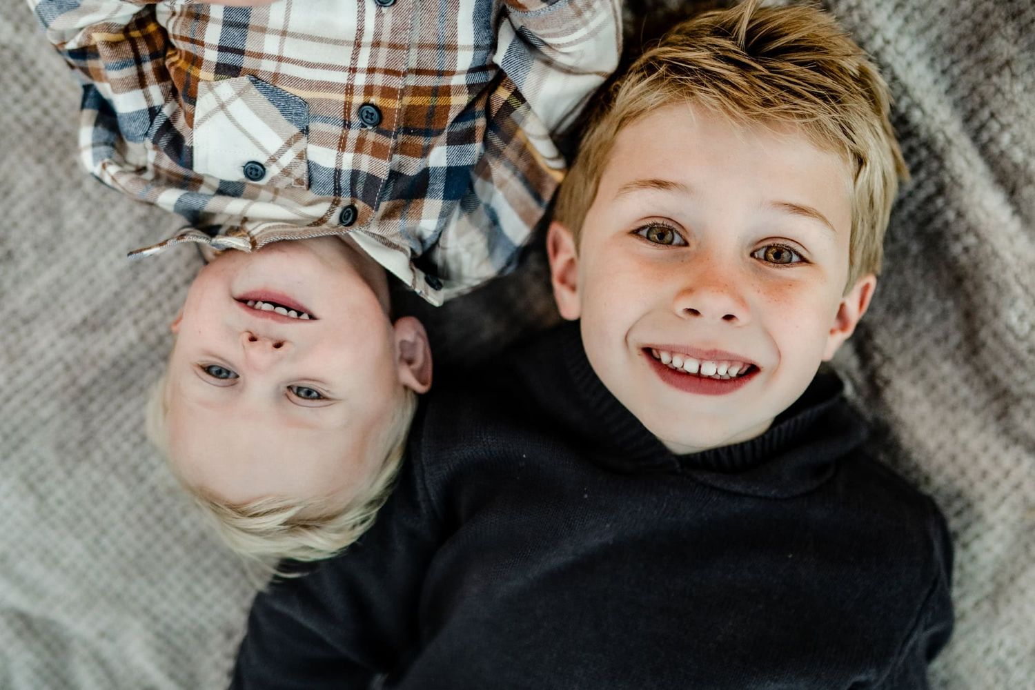 Two children lying on a gray blanket smiling at the camera from above.