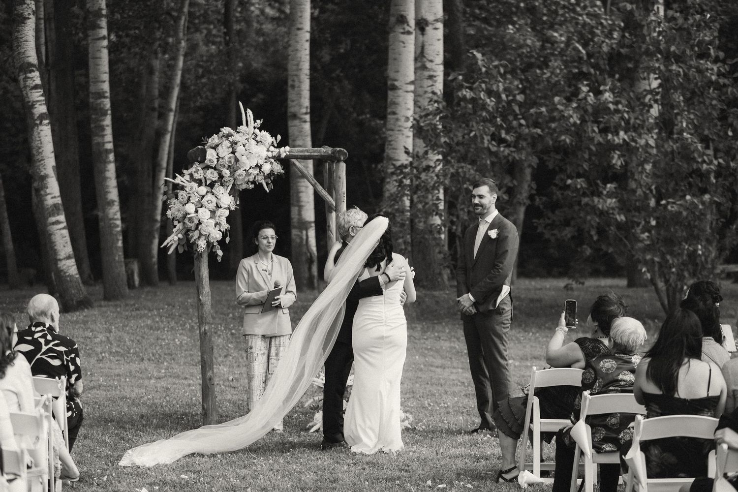 A black and white wedding photograph showing people gathered in a forest setting for an outdoor ceremony.