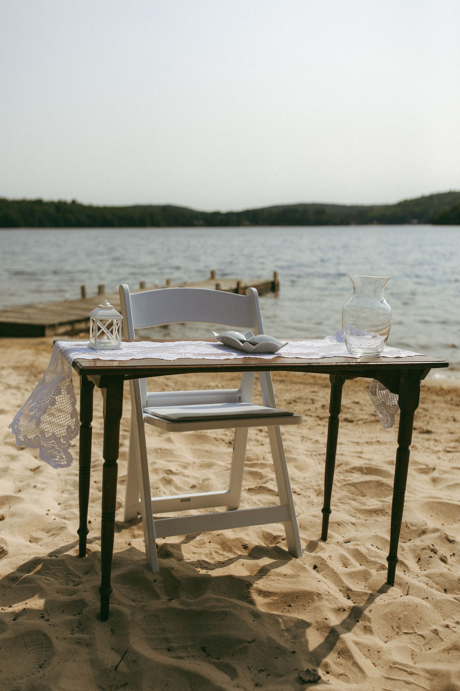 A rustic wooden table with chairs sits on a sandy beach overlooking a misty lake or ocean view.