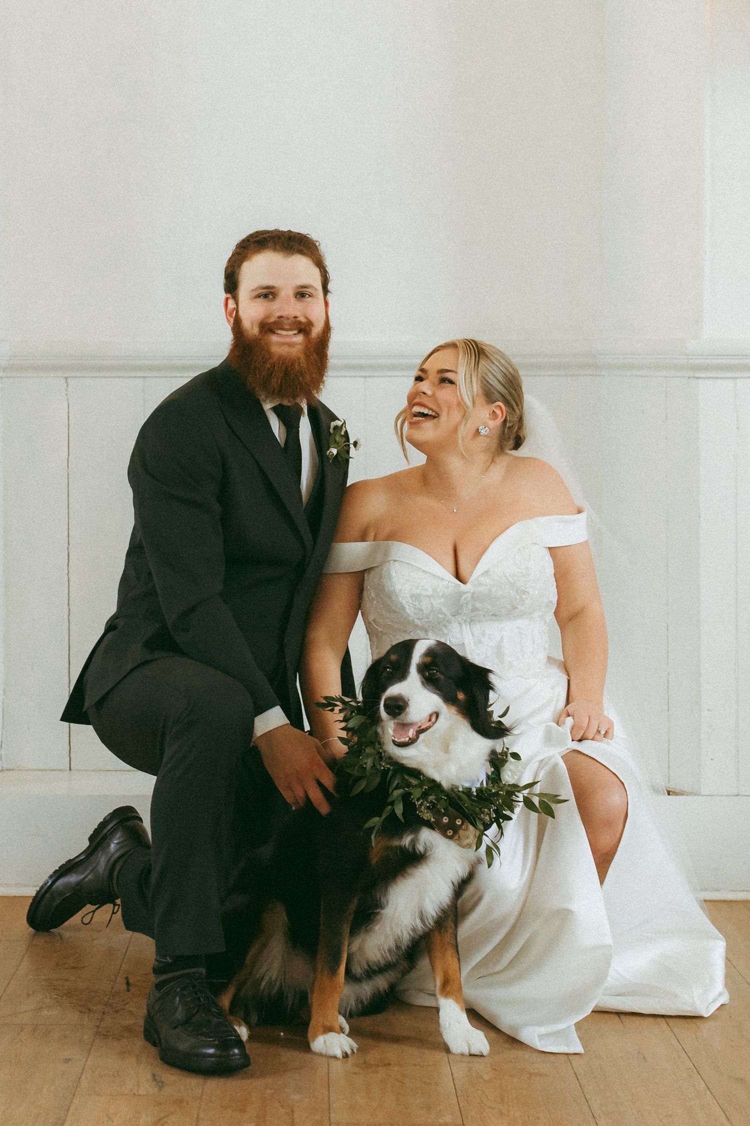 A happy couple in wedding attire sits with their Border Collie dog who wears a floral collar on their special day.