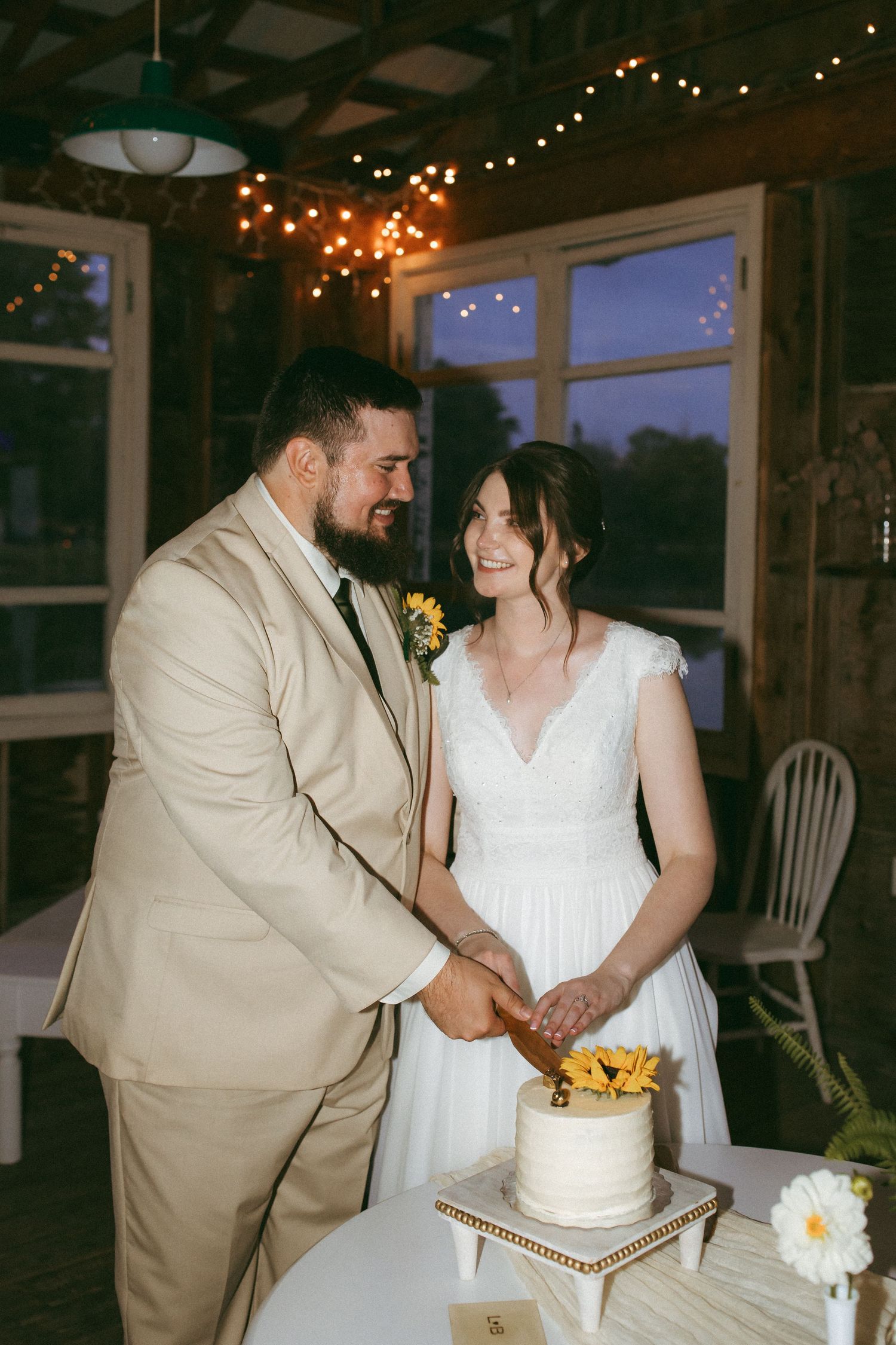 A couple shares intimate moments while cutting their wedding cake together in a rustic venue decorated with string lights.