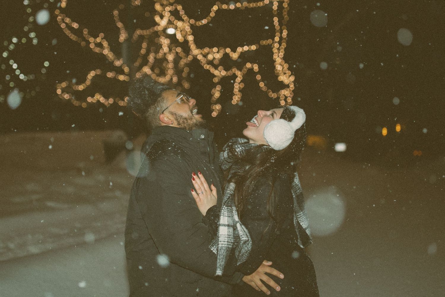 A couple stands together outdoors at night while snow falls around them with glowing string lights in the background.