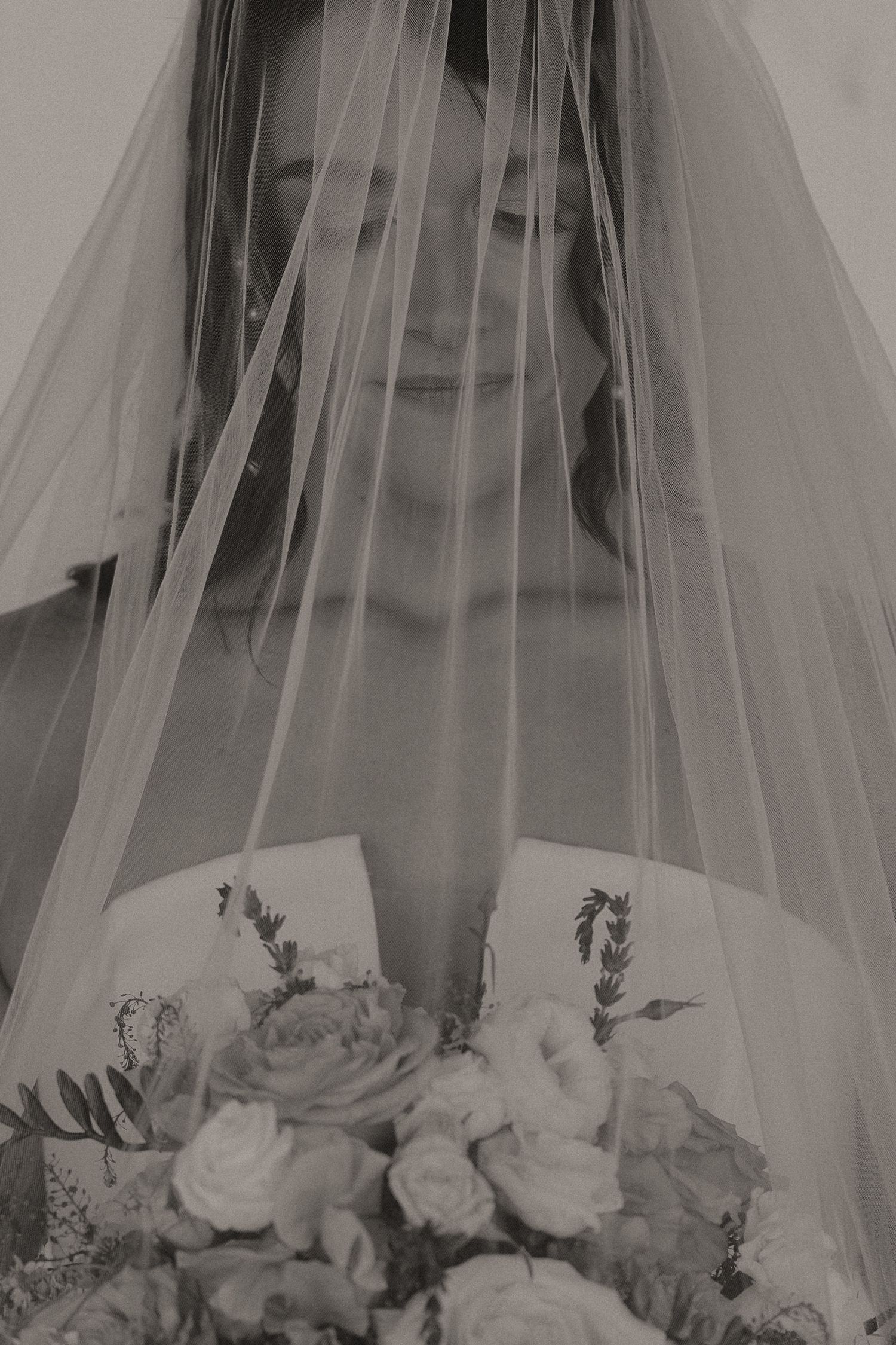 Soft black and white portrait of a smiling bride holding a bouquet, her face partially covered by a veil.