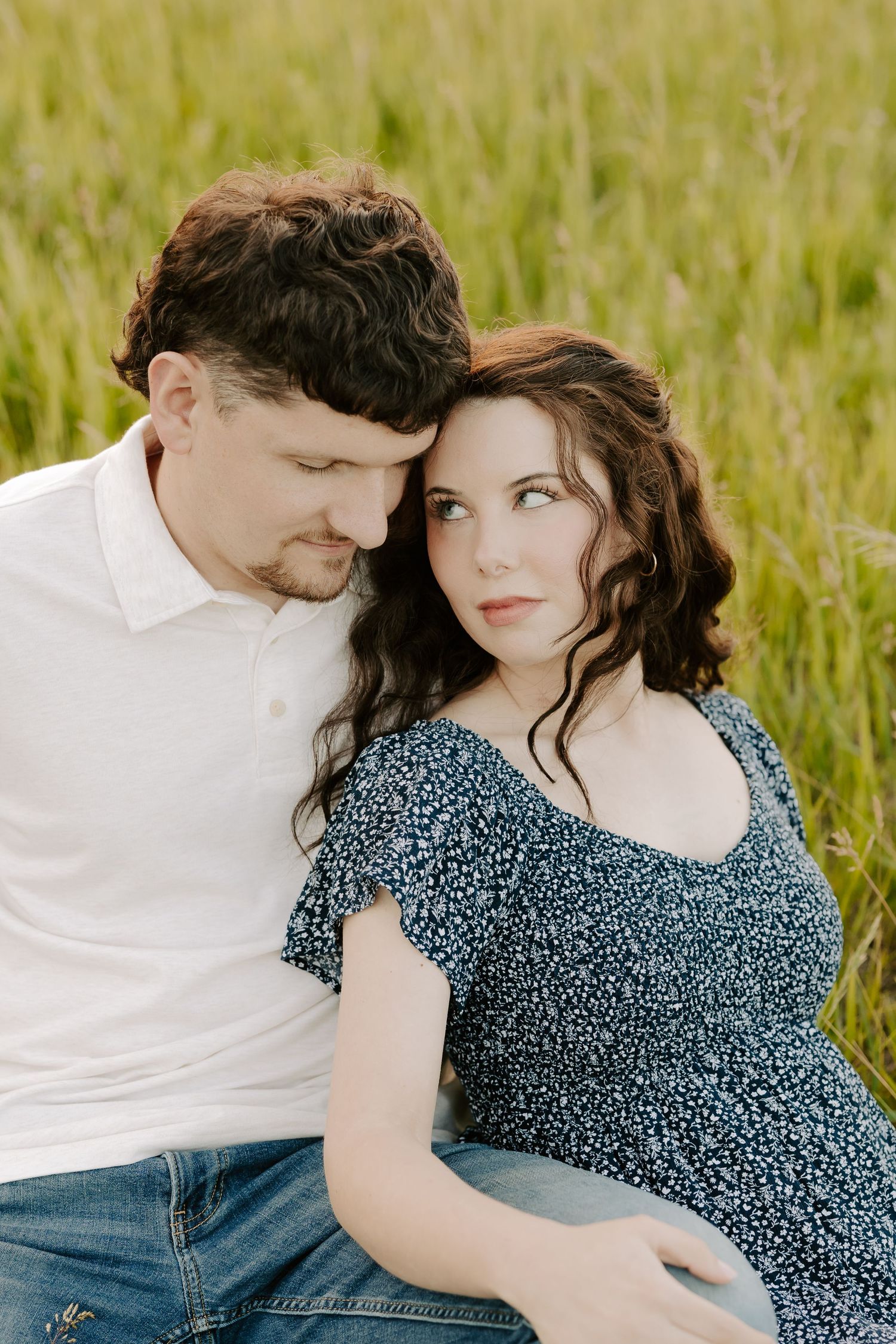 A romantic couple sits together in a green field, leaning close with tender expressions in soft sunlight.