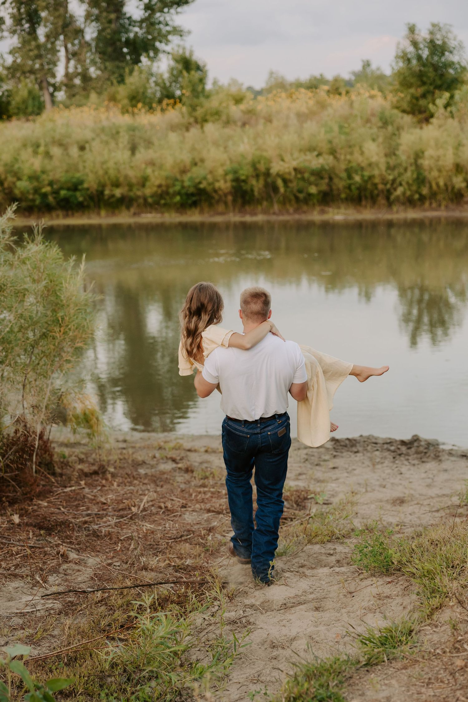 A man carries a woman in his arms along a sandy riverbank surrounded by lush autumn foliage.