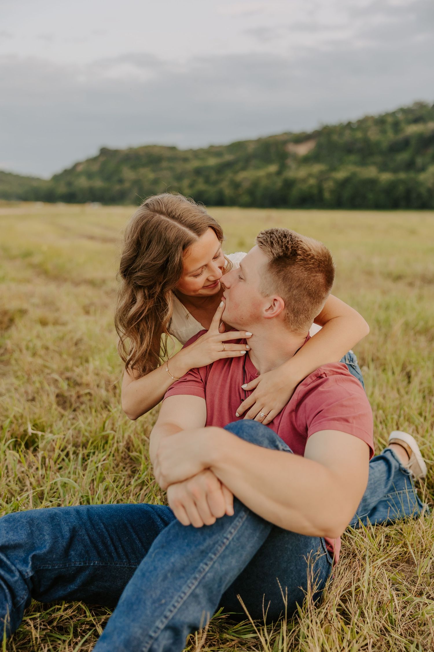 Happy couple sharing a playful kiss while sitting together in a lush green meadow with rolling hills.