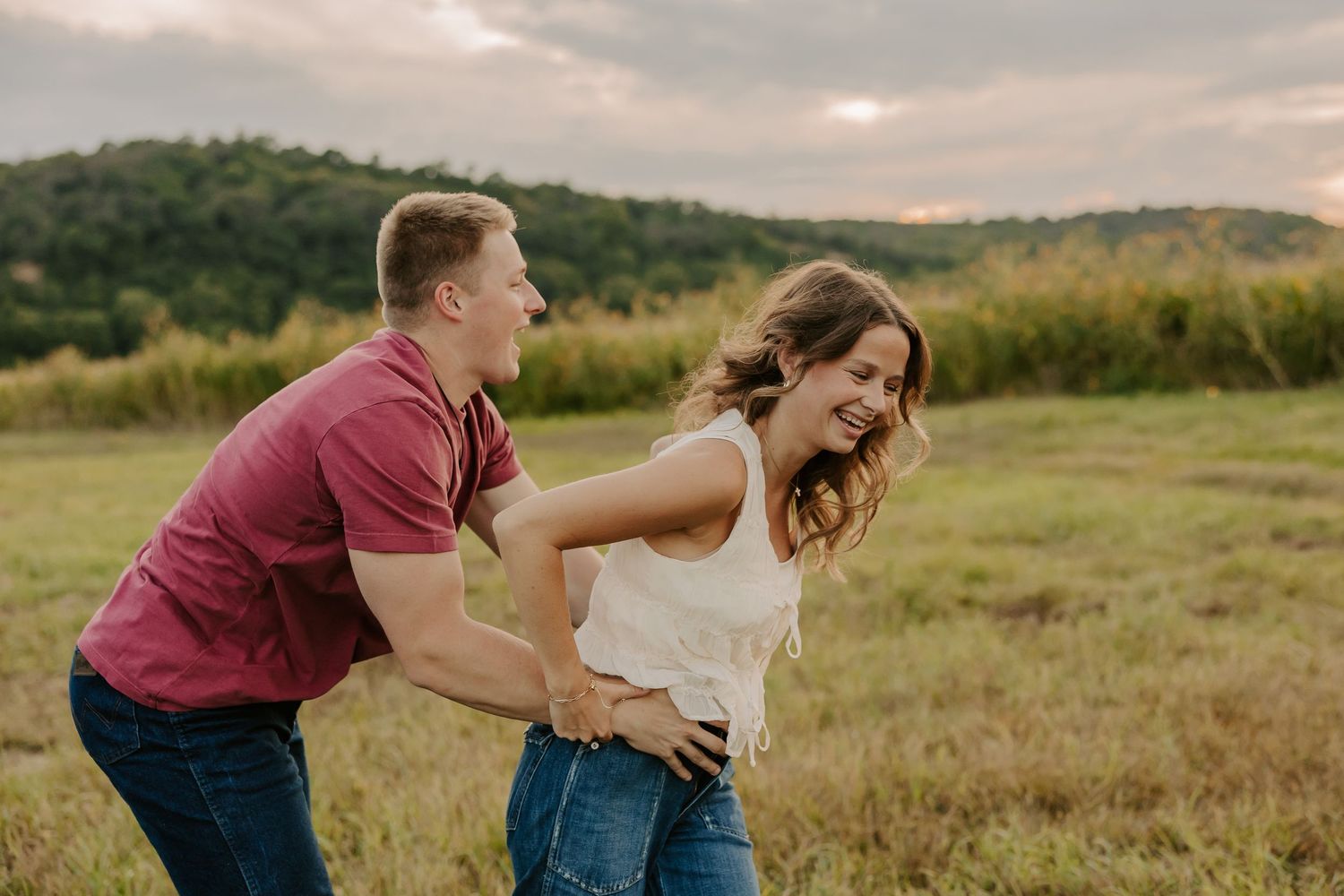 A joyful couple laughs and plays in a green meadow with scenic hills under a dramatic cloudy sky.
